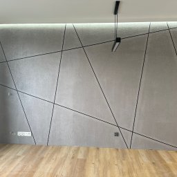 Usługi minikoparką Edengard Andrzej Folwarski - Szara ściana z dekoracyjną fakturą i czarnymi, ukośnymi listwami tworzącymi geometryczny wzór, oświetlona od góry taśmą LED, z widocznymi gniazdkami elektrycznymi i wiszącą, nowoczesną lampą.