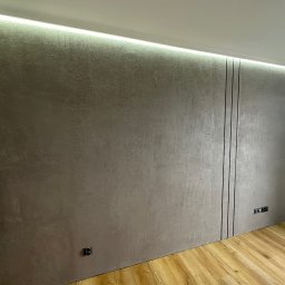 Usługi minikoparką Edengard Andrzej Folwarski - Ściana w pokoju z szarą, strukturalną powierzchnią, oświetleniem LED wzdłuż sufitu i trzema pionowymi czarnymi liniami dekoracyjnymi, widoczne gniazdka elektryczne.