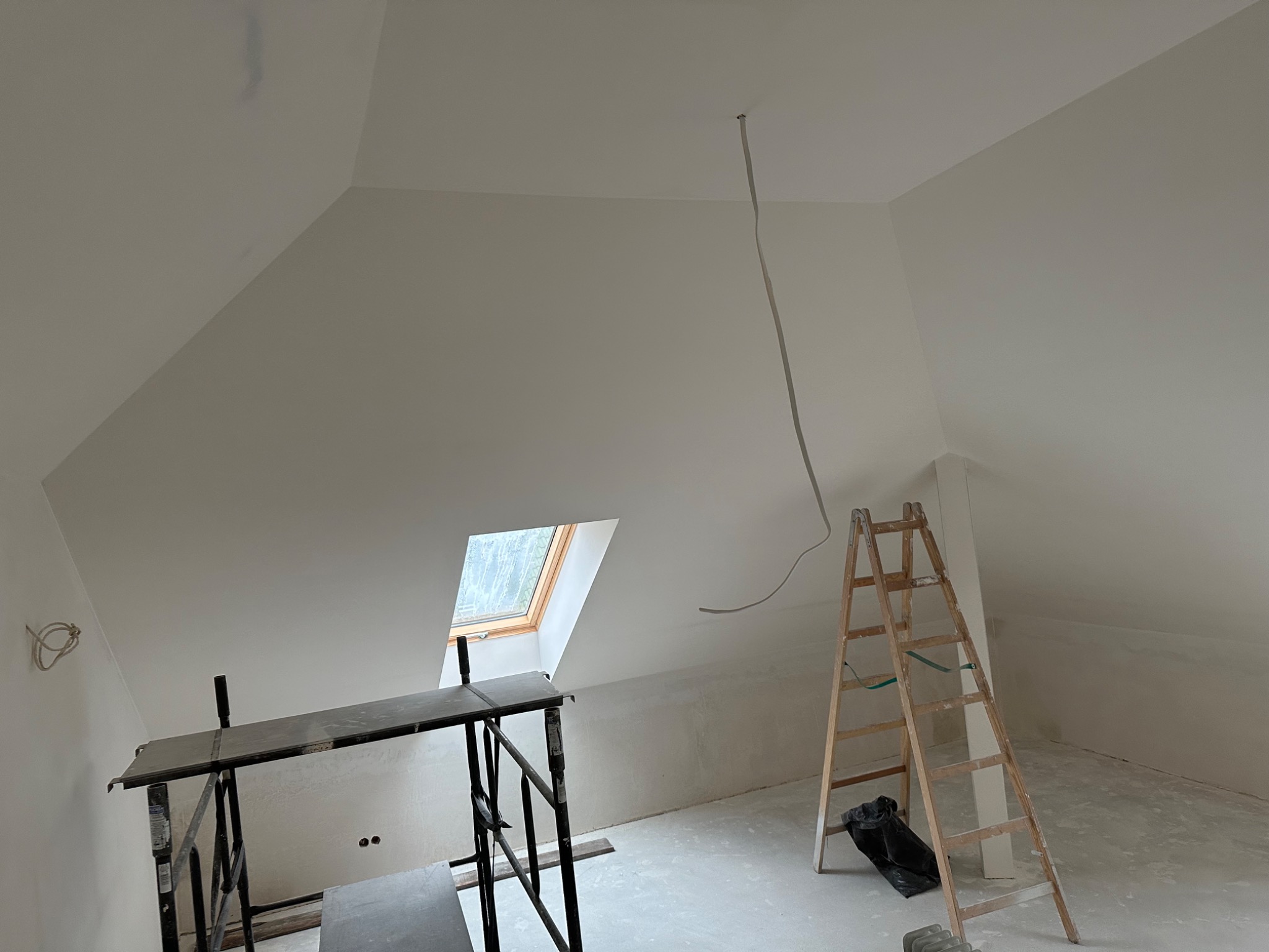 Poddasze po pracach wykończeniowych, widoczne rusztowanie, drabina i kabel zwisający z sufitu, okno dachowe w tle.