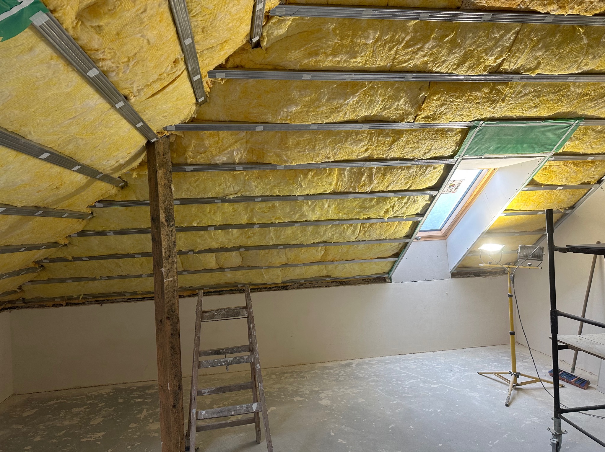 Poddasze w trakcie izolacji termicznej, widoczne żółte ocieplenie między metalowymi profilami, okno dachowe z zieloną osłoną, drewniany słup i drabina, lampa budowlana na statywie.