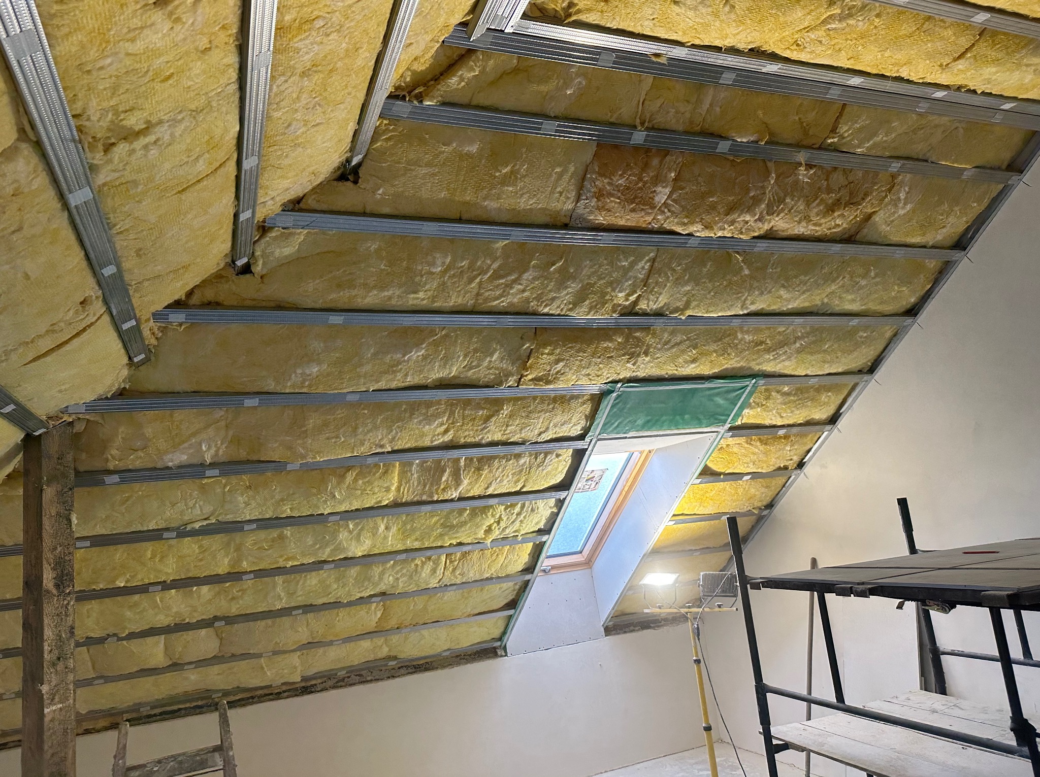 Poddasze w trakcie prac izolacyjnych, widoczna wełna mineralna między metalowymi profilami, okno dachowe z folią ochronną, rusztowanie w tle.