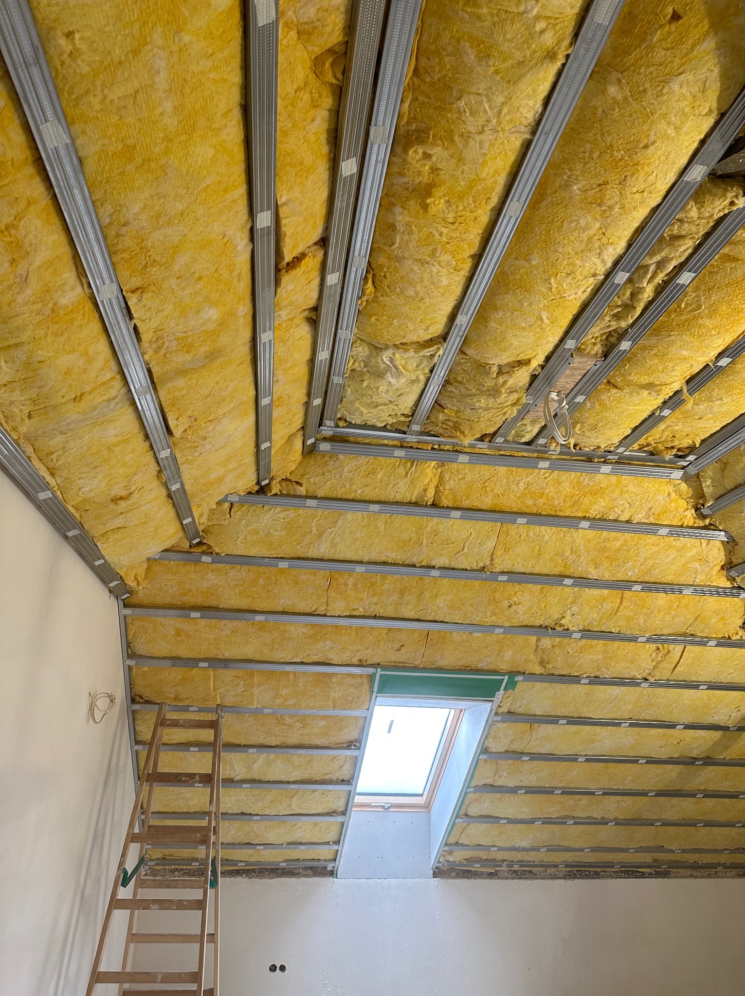 Żółta wełna mineralna izolująca skośny dach poddasza, zamontowana między metalowymi profilami, widoczne okno dachowe i drabina oparta o ścianę.