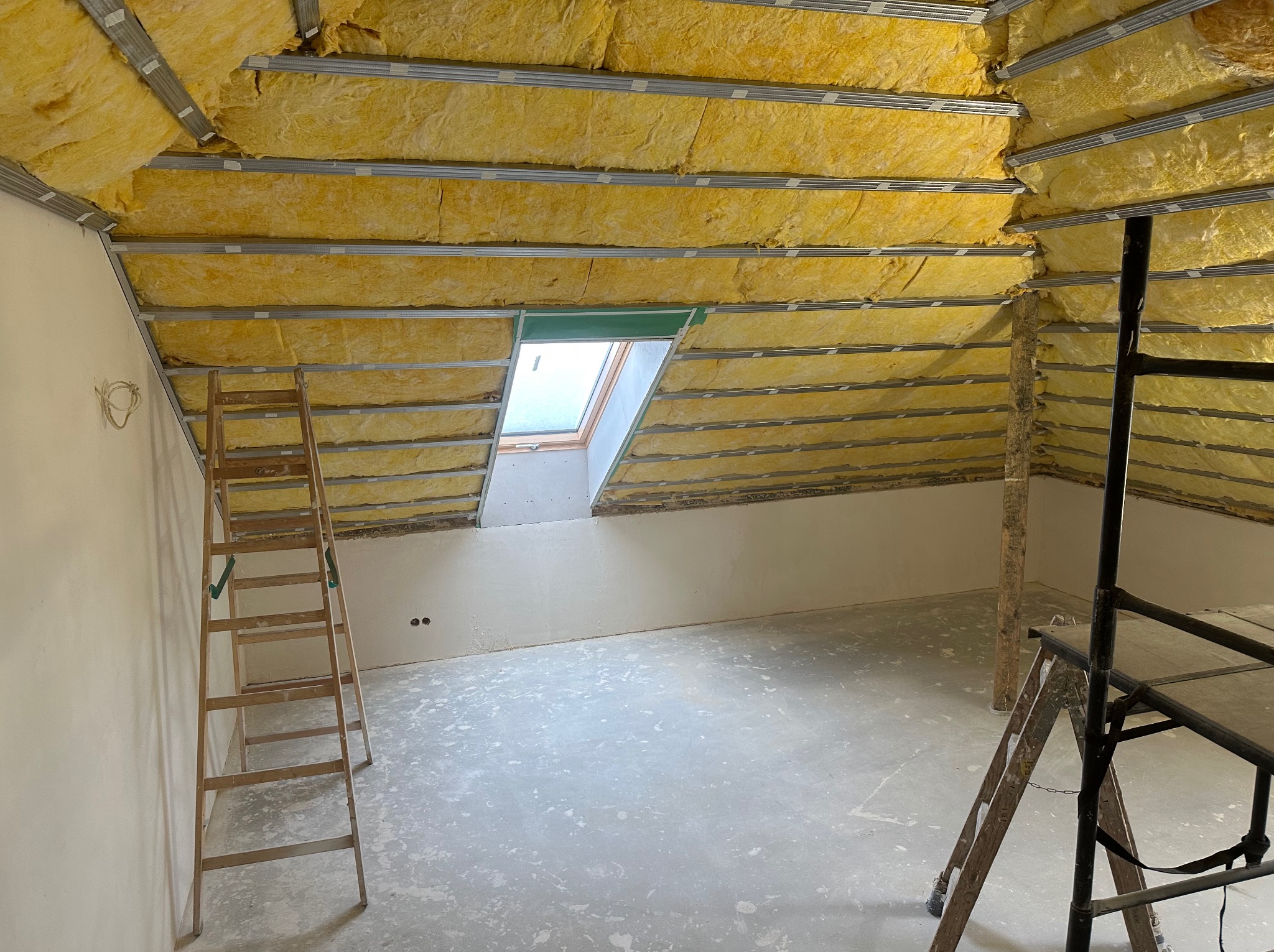 Poddasze w trakcie izolacji termicznej, widoczne żółte panele wełny mineralnej między metalowymi profilami, okno dachowe, drabina drewniana i rusztowanie.