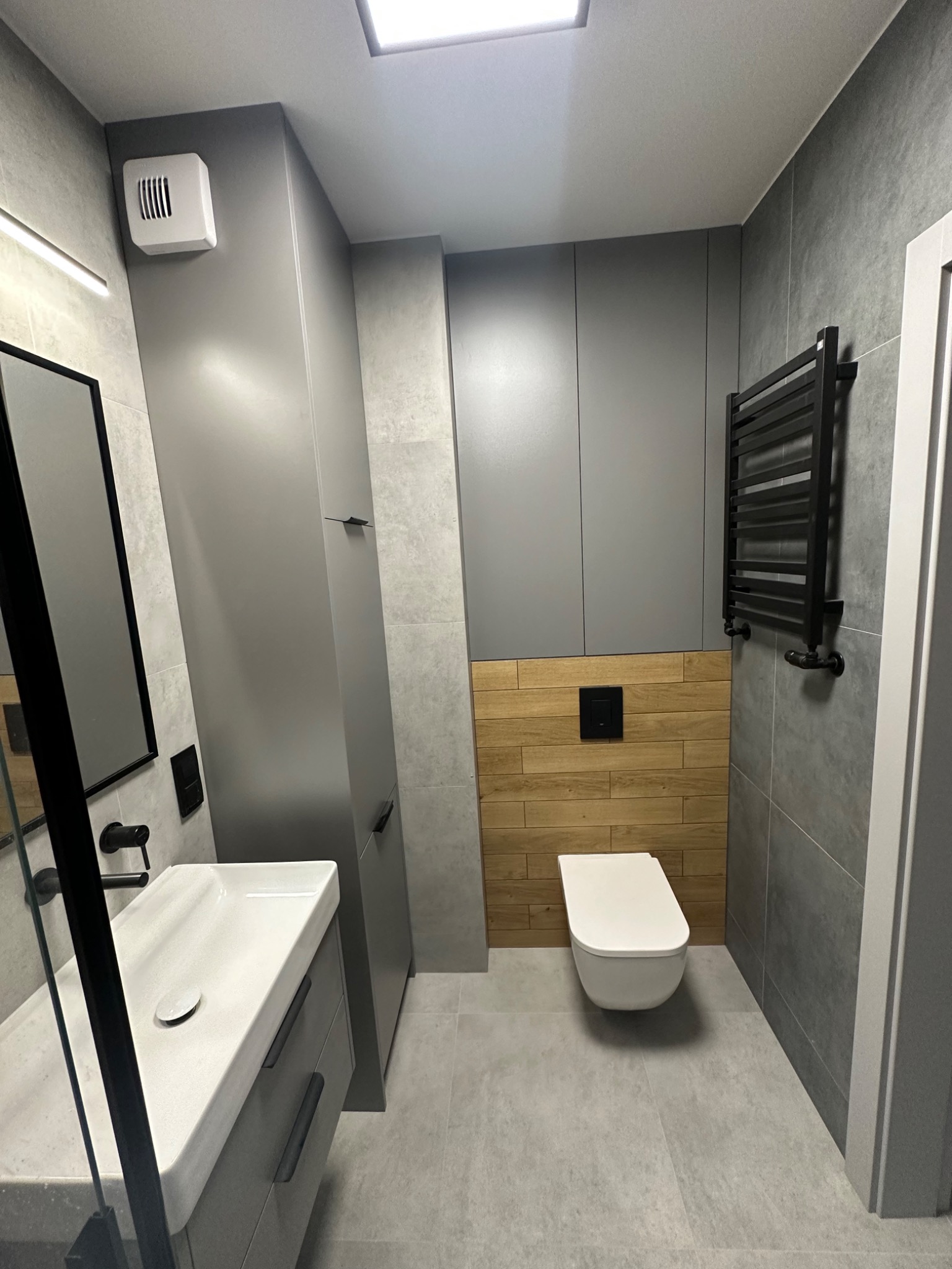 Nowoczesna łazienka w szarościach z drewnianym akcentem: wisząca toaleta, umywalka z szafką, grzejnik drabinkowy i płytki imitujące kamień.