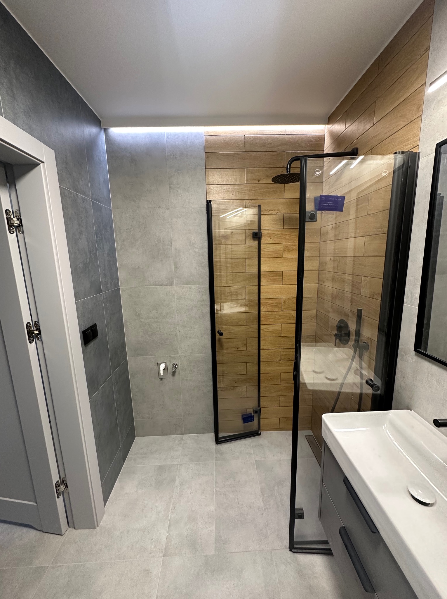 Nowoczesna łazienka z kabiną prysznicową typu walk-in z czarnymi ramami, drewnopodobnymi płytkami na ścianie prysznica, szarą ceramiką i podświetleniem LED sufitu.