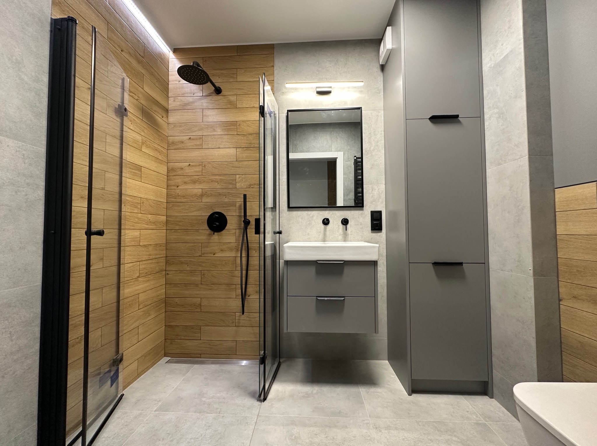 Nowoczesna łazienka z kabiną prysznicową typu walk-in z drewnopodobnymi panelami, szarą szafką z umywalką i wysoką szafą, wykończona szarymi płytkami.