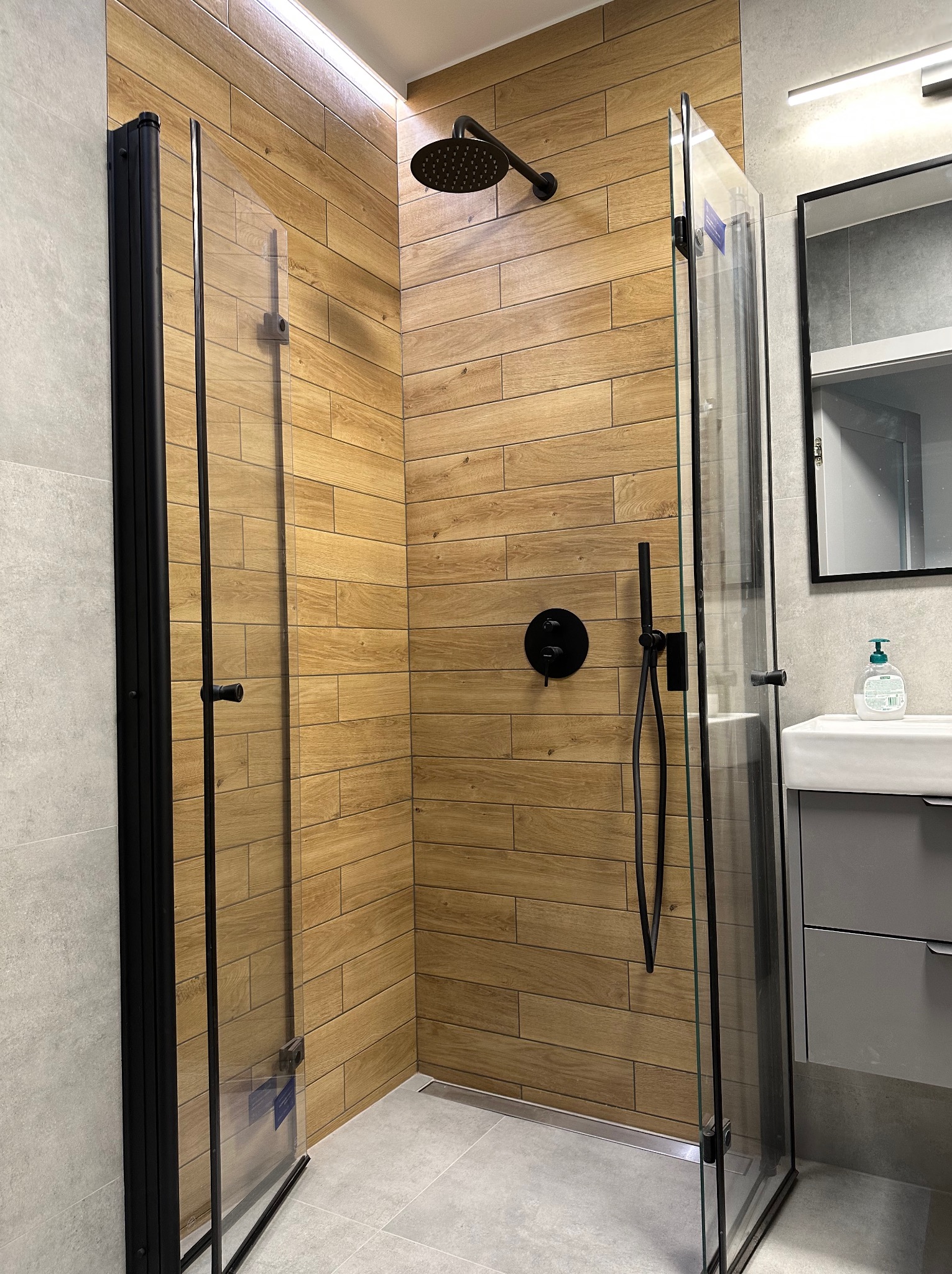Nowoczesna kabina prysznicowa z czarnymi elementami i ścianami wyłożonymi płytkami imitującymi drewno, widoczna umywalka z szafką i lustrem.