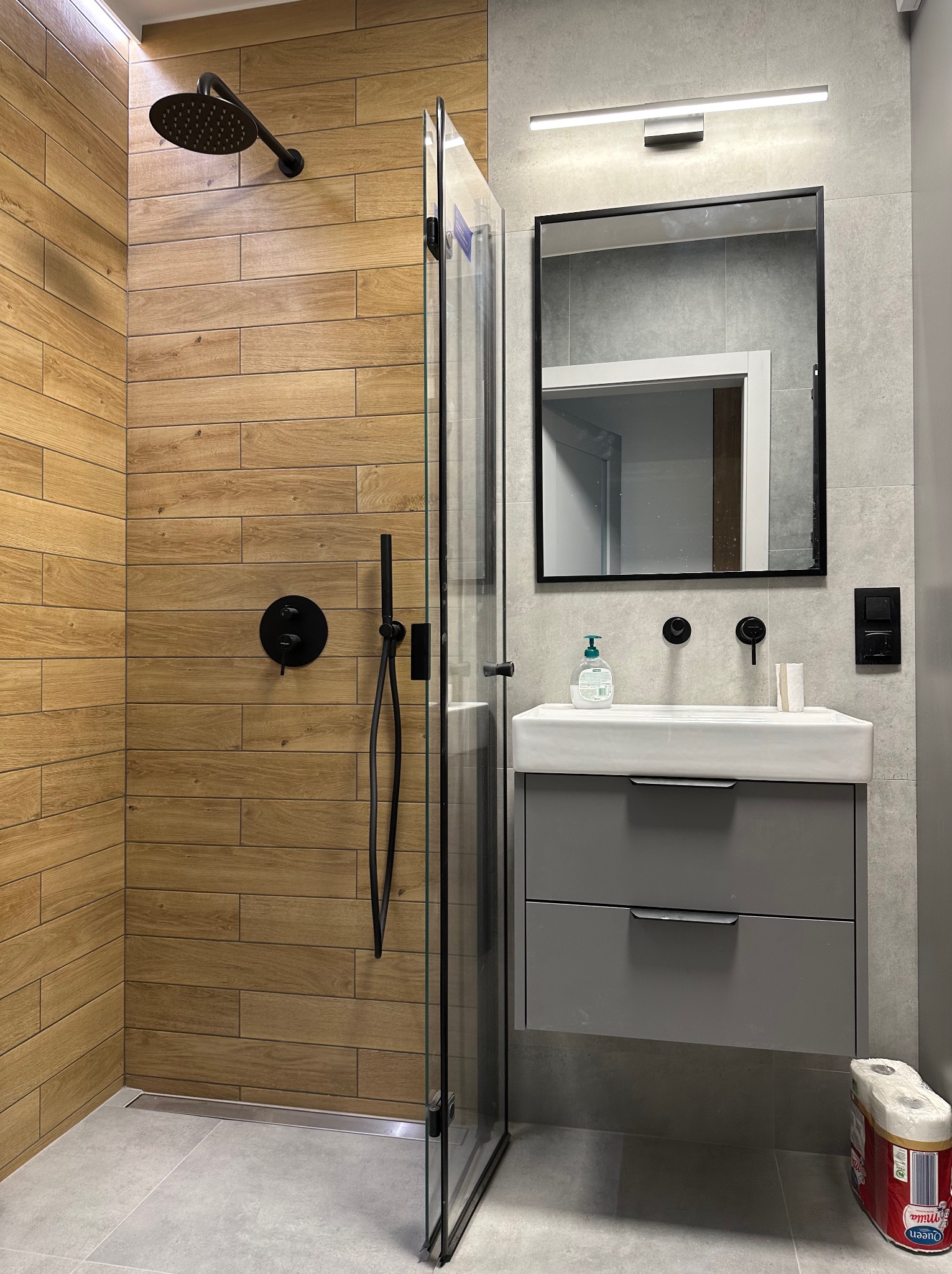 Nowoczesna łazienka z drewnopodobnymi płytkami w strefie prysznica, czarną armaturą i szarą szafką pod umywalką, minimalistyczny design.