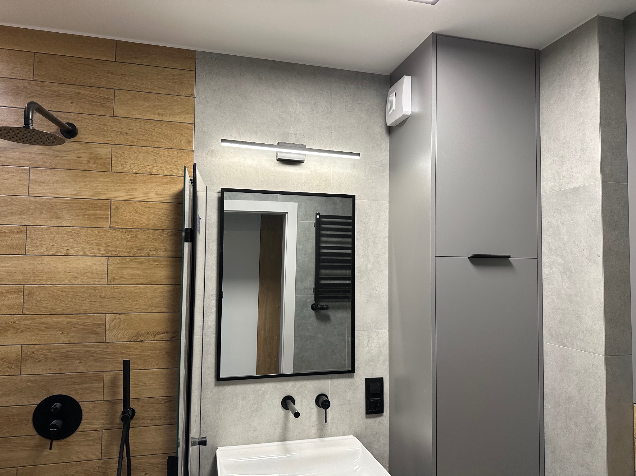Nowoczesna łazienka z drewnopodobną okładziną ścienną, czarną armaturą i minimalistycznym oświetleniem LED nad lustrem.