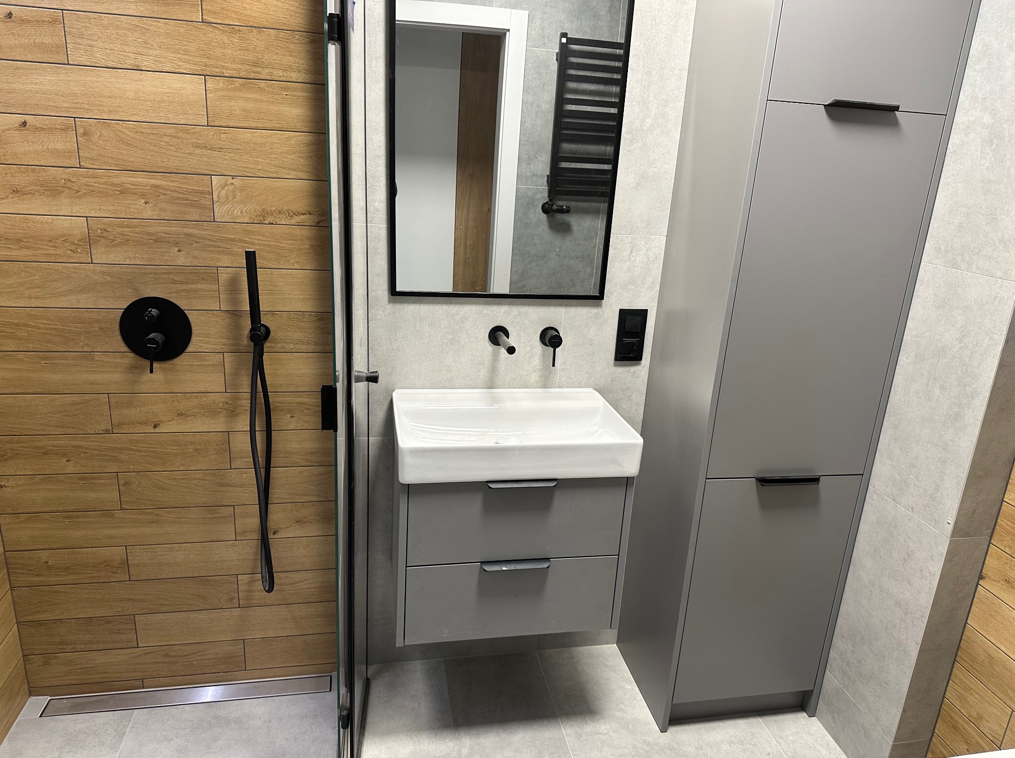 Nowoczesna łazienka z drewnopodobnymi płytkami ściennymi w strefie prysznica, szarą szafką pod umywalką i wysoką szafką, czarną armaturą oraz lustrem w czarnej ramie.