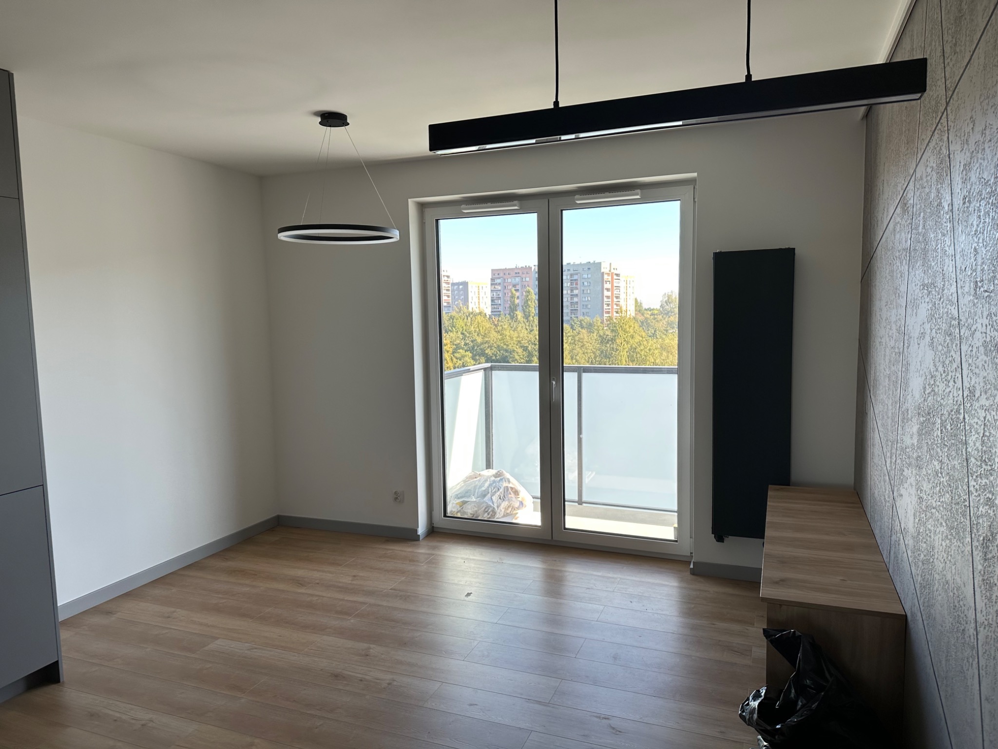 Pusty pokój z drewnianą podłogą i balkonem, widok na budynki w tle, nowoczesne oświetlenie sufitowe, ściana z dekoracyjną fakturą.