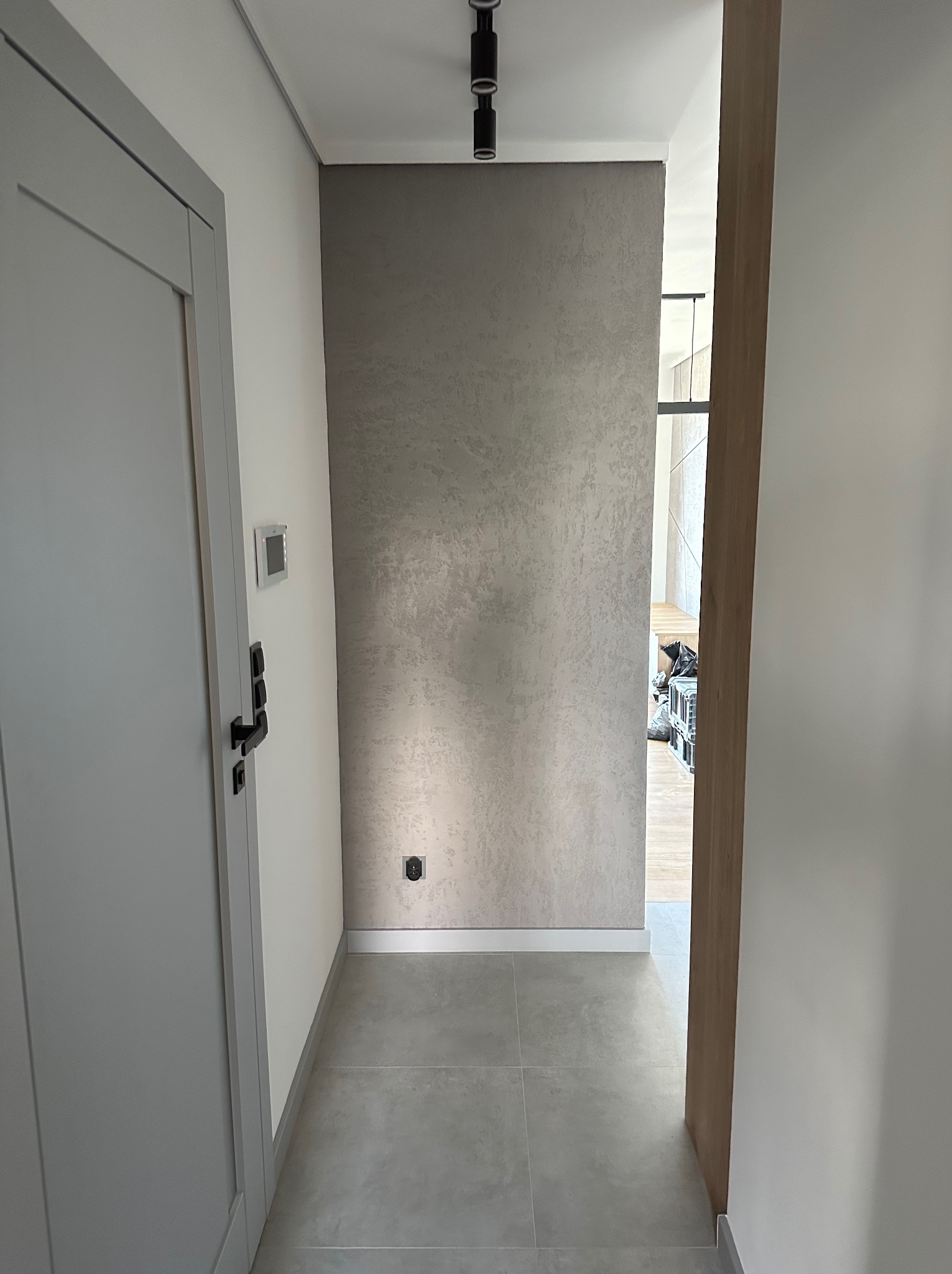 Korytarz z szarą ścianą o teksturowanej powierzchni, szarą podłogą z płytek i szarymi drzwiami po lewej stronie; widoczny fragment wnętrza przez otwarte drzwi.