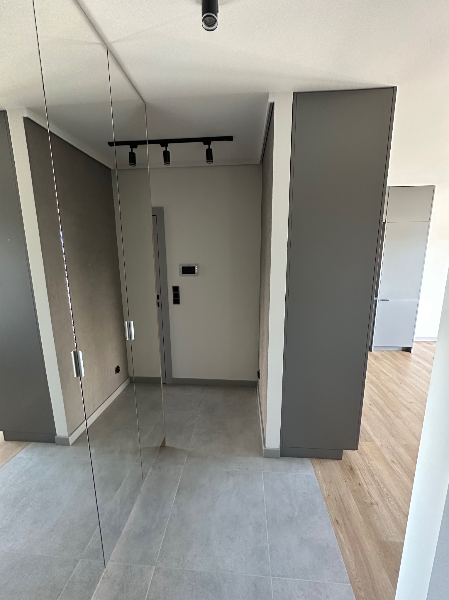Nowoczesny przedpokój z lustrzaną szafą w zabudowie, szara płytka podłogowa i drewniana podłoga w dalszej części mieszkania, oświetlenie szynowe na suficie.