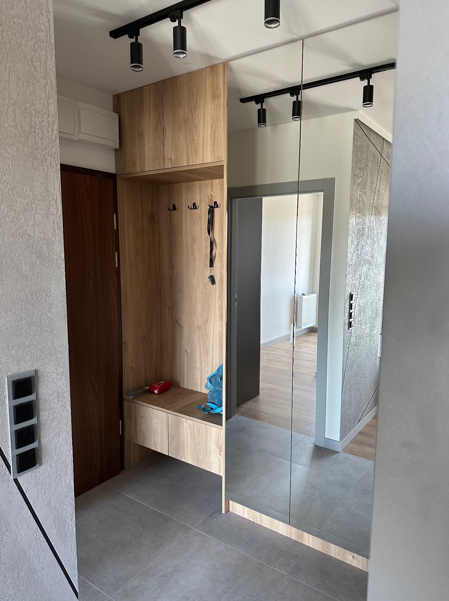 Nowoczesny przedpokój z drewnianą zabudową, wbudowanym siedziskiem, lustrem na ścianie, szarą podłogą i oświetleniem punktowym na suficie.