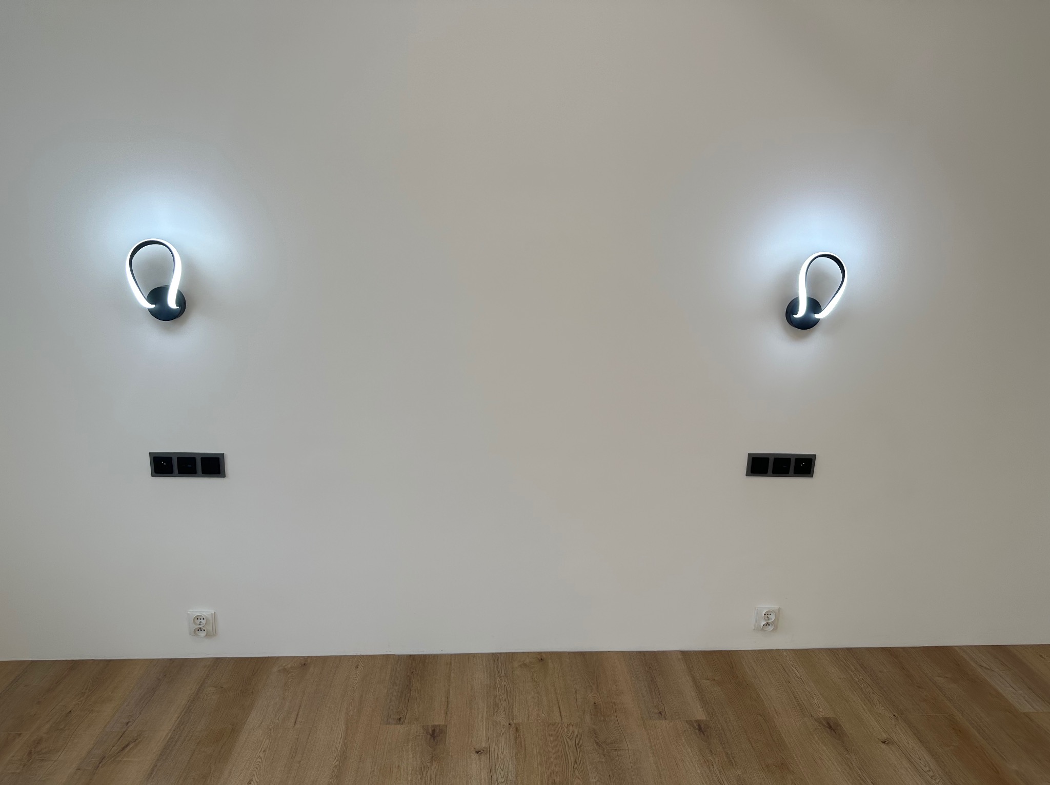 Biała ściana z dwoma nowoczesnymi, świecącymi kinkietami o opływowym kształcie, symetrycznie rozmieszczonymi nad czarnymi gniazdkami elektrycznymi i jasnobrązową podłogą z paneli.