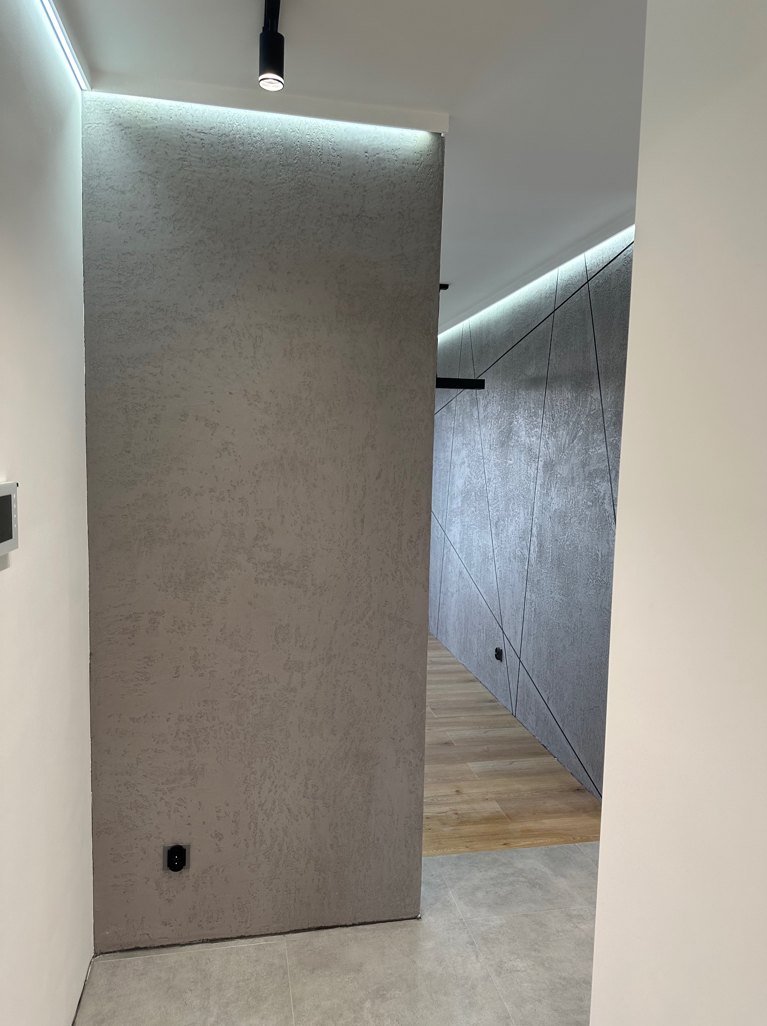 Nowoczesny korytarz z dekoracyjną ścianą pokrytą szarą, strukturalną gładzią, oświetleniem LED i podłogą z płytek imitujących drewno.