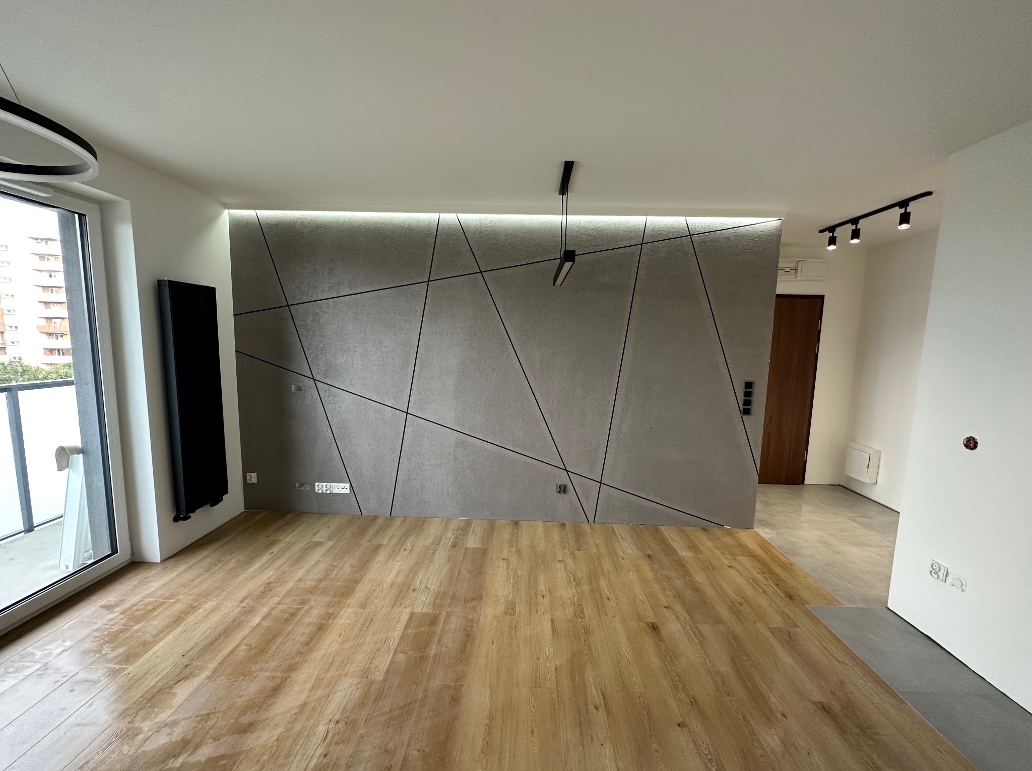 Wykończone wnętrze w Rupniowie: Ściana z dekoracyjną szarą strukturą i geometrycznym wzorem, podświetlona listwą LED, drewniana podłoga, nowoczesne oświetlenie sufitowe.