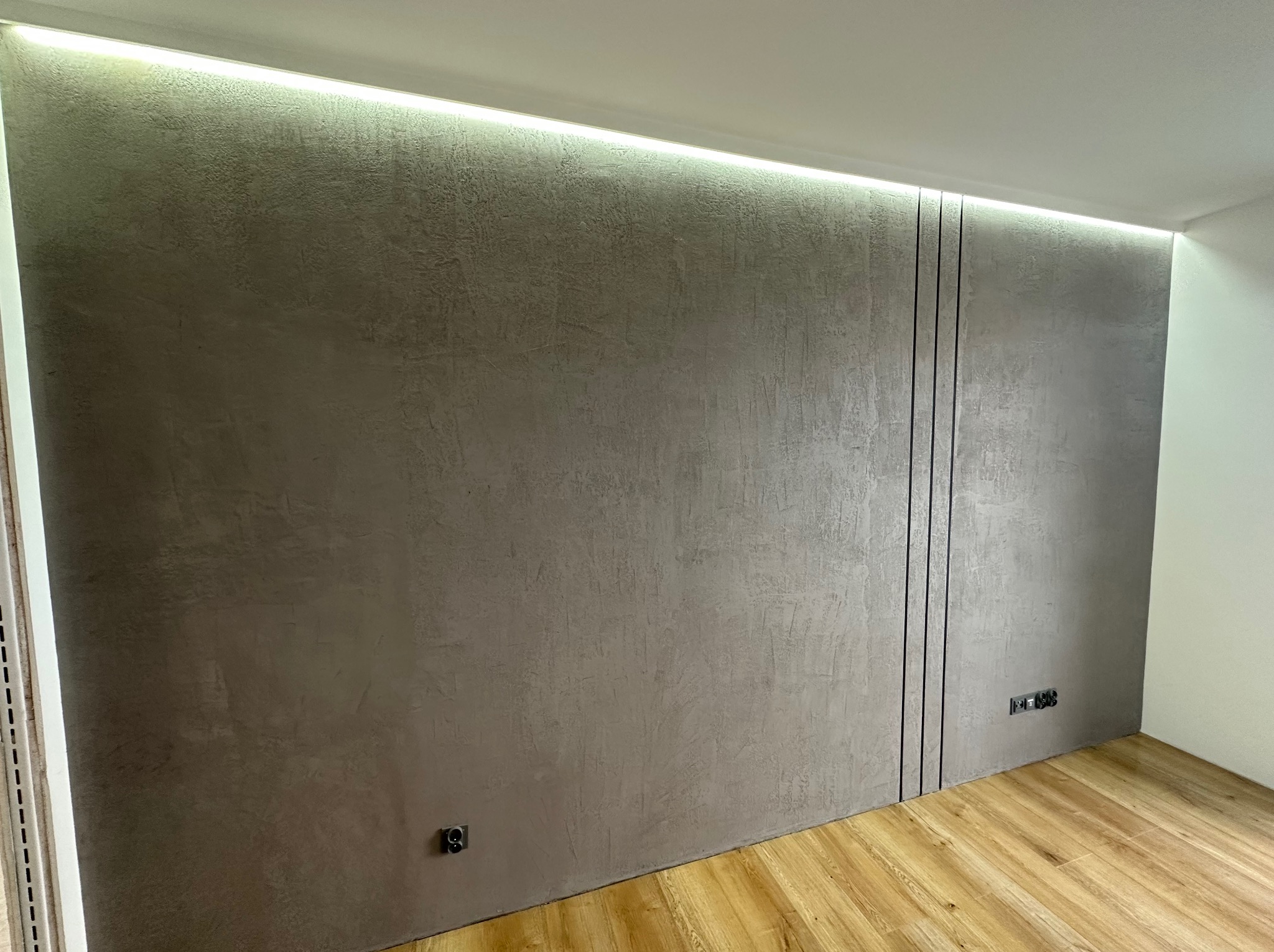 Ściana w pokoju z szarą, strukturalną powierzchnią, oświetleniem LED wzdłuż sufitu i trzema pionowymi czarnymi liniami dekoracyjnymi, widoczne gniazdka elektryczne.