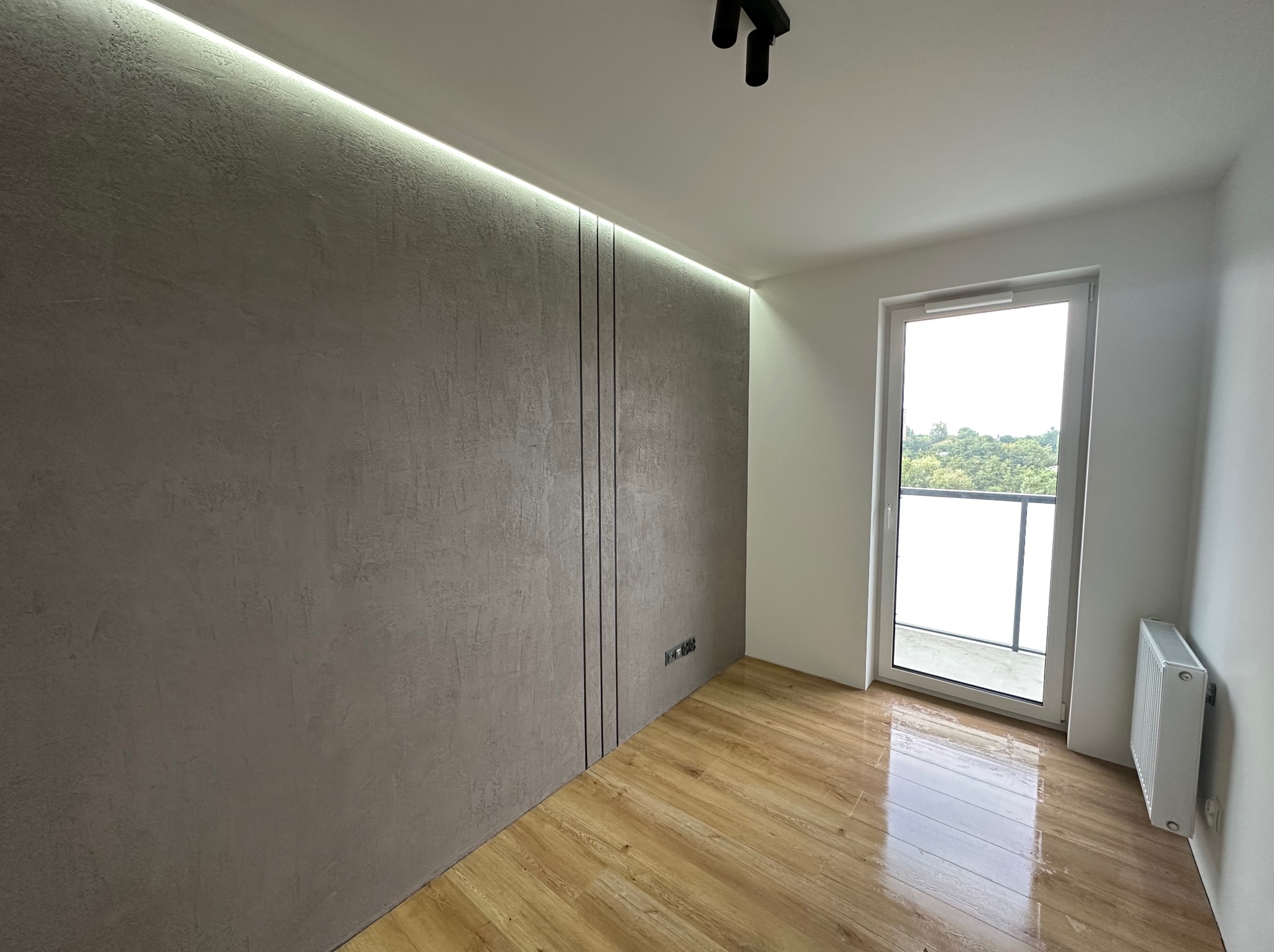 Pusty pokój po remoncie z drewnianą podłogą, ścianą pokrytą dekoracyjną szarą fakturą z pionowymi wstawkami i oświetleniem LED, białe okno balkonowe i grzejnik.