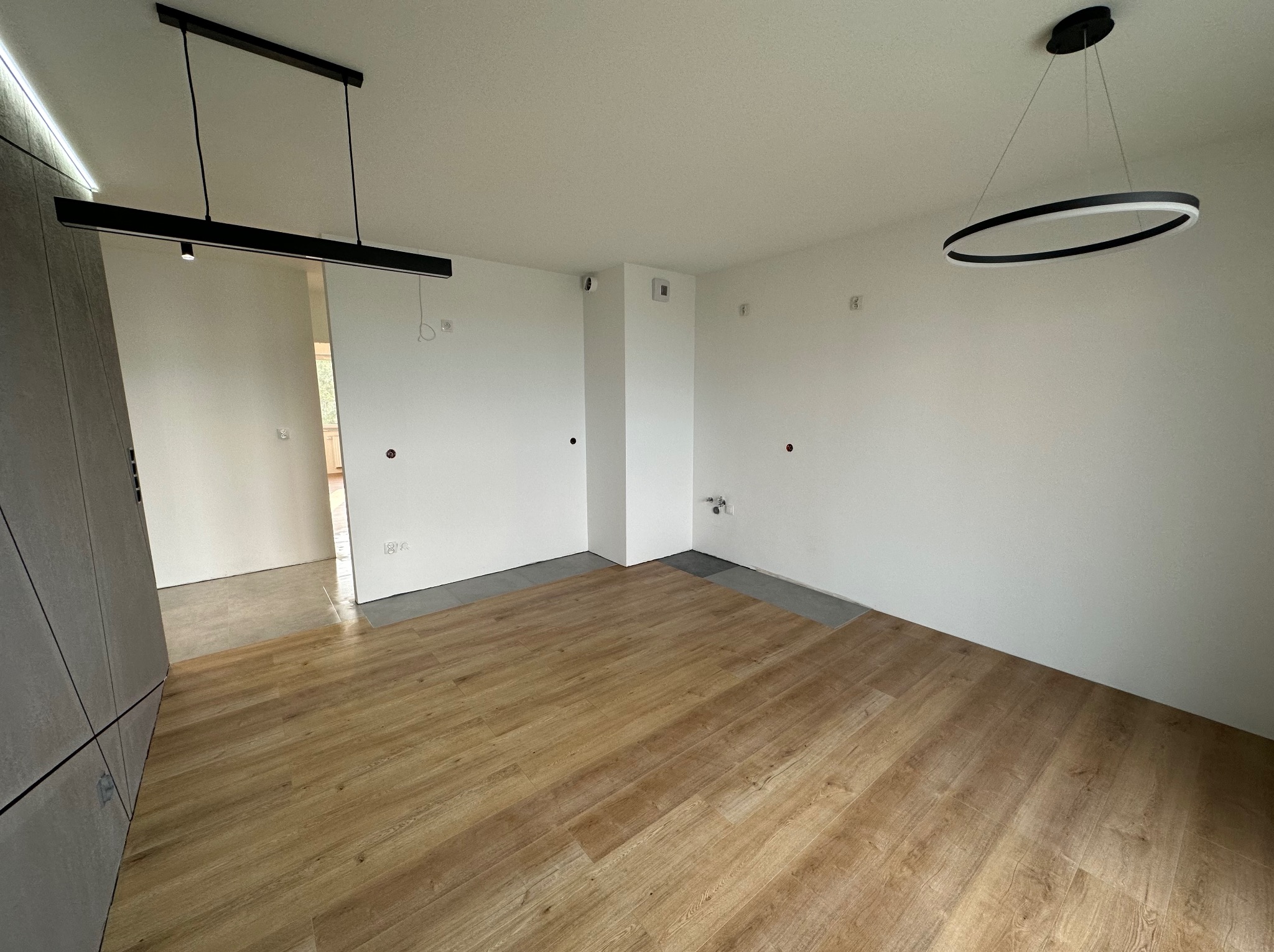 Wnętrze pokoju po remoncie z drewnianą podłogą, białymi ścianami z widocznymi puszkami elektrycznymi i dwoma nowoczesnymi lampami sufitowymi: prostokątną i okrągłą.