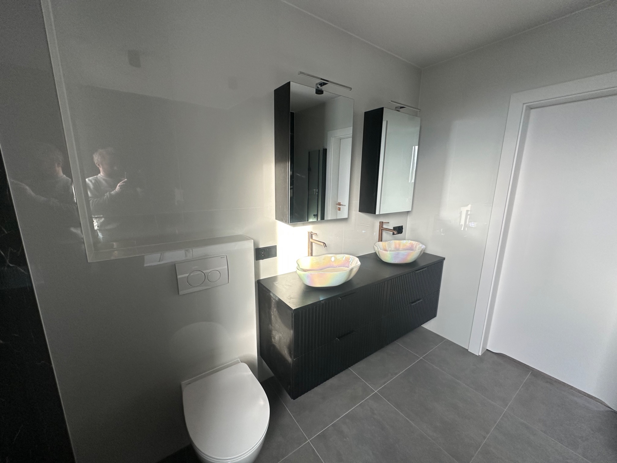 Nowoczesna łazienka z podwójną umywalką w odcieniach szarości i bieli, z minimalistyczną toaletą podwieszaną i lustrami z oświetleniem. Widoczne płytki podłogowe i gładkie ściany.