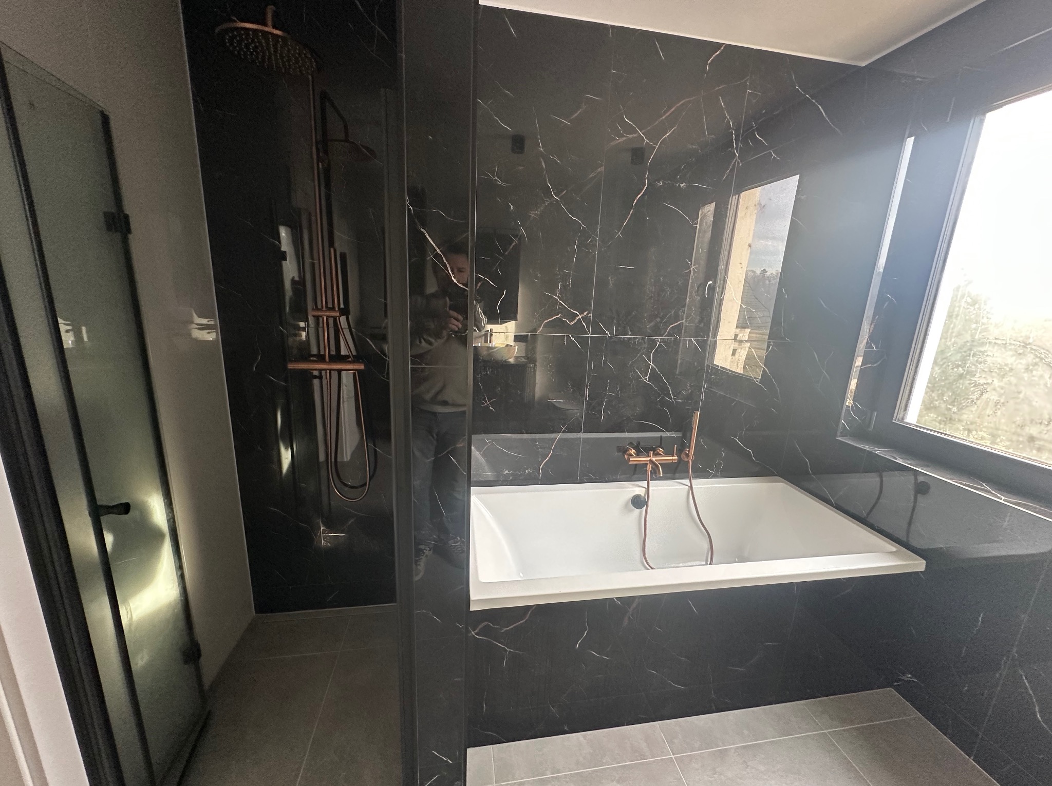 Nowoczesna łazienka z czarnymi płytkami imitującymi marmur, białą wanną i miedzianą armaturą. Widoczna kabina prysznicowa i okno z widokiem na zewnątrz.