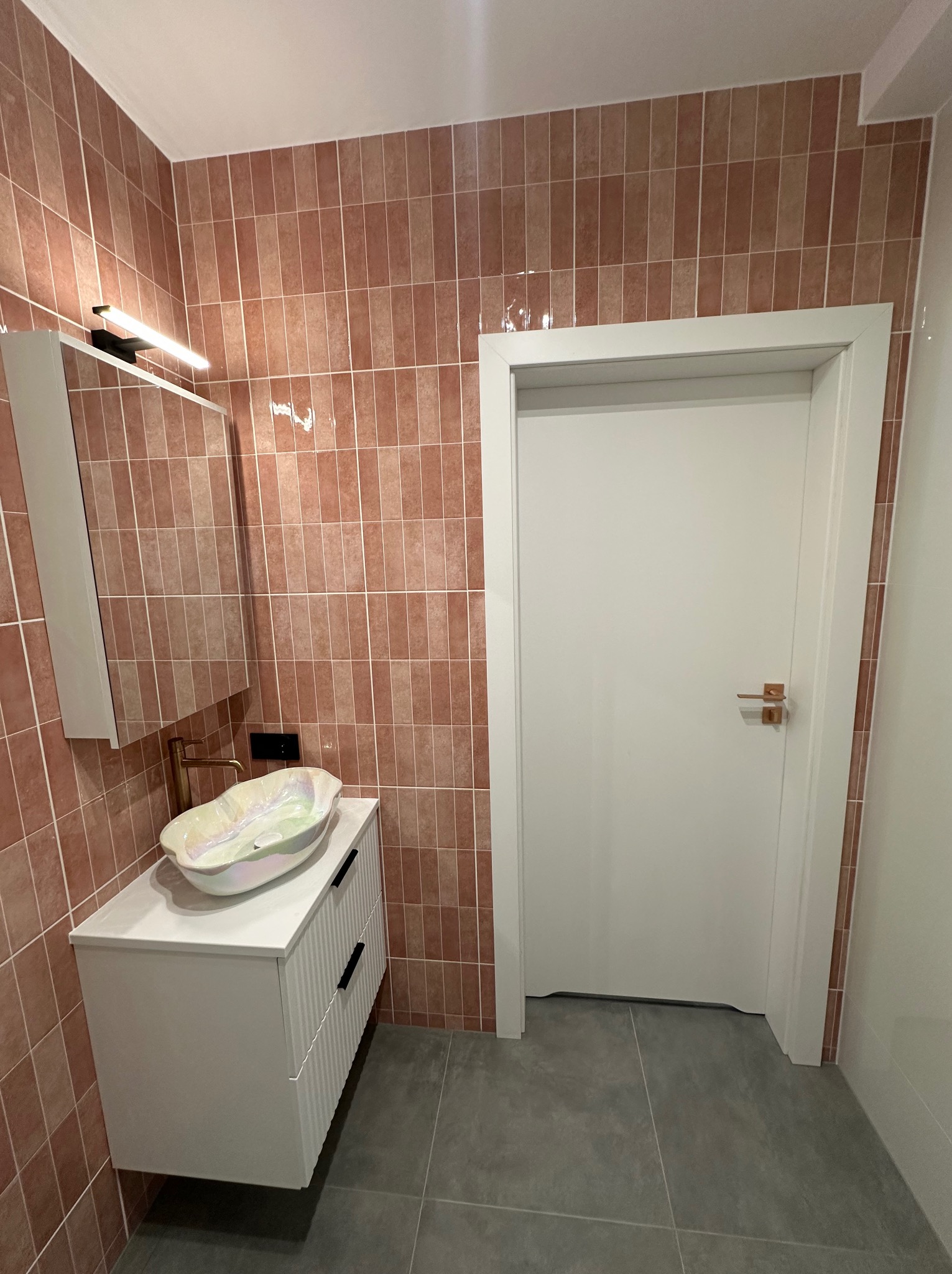 Wykończona łazienka z umywalką na szafce, brązowe płytki na ścianach i szara podłoga. Białe drzwi z złotą klamką. Nowoczesne oświetlenie nad lustrem.