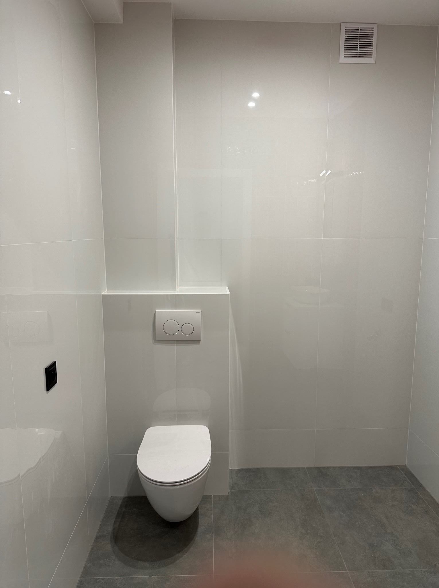 Nowoczesna toaleta w minimalistycznym stylu: białe płytki ścienne z szarą podłogą. Wisząca muszla klozetowa i przycisk spłukujący. Widoczna kratka wentylacyjna.