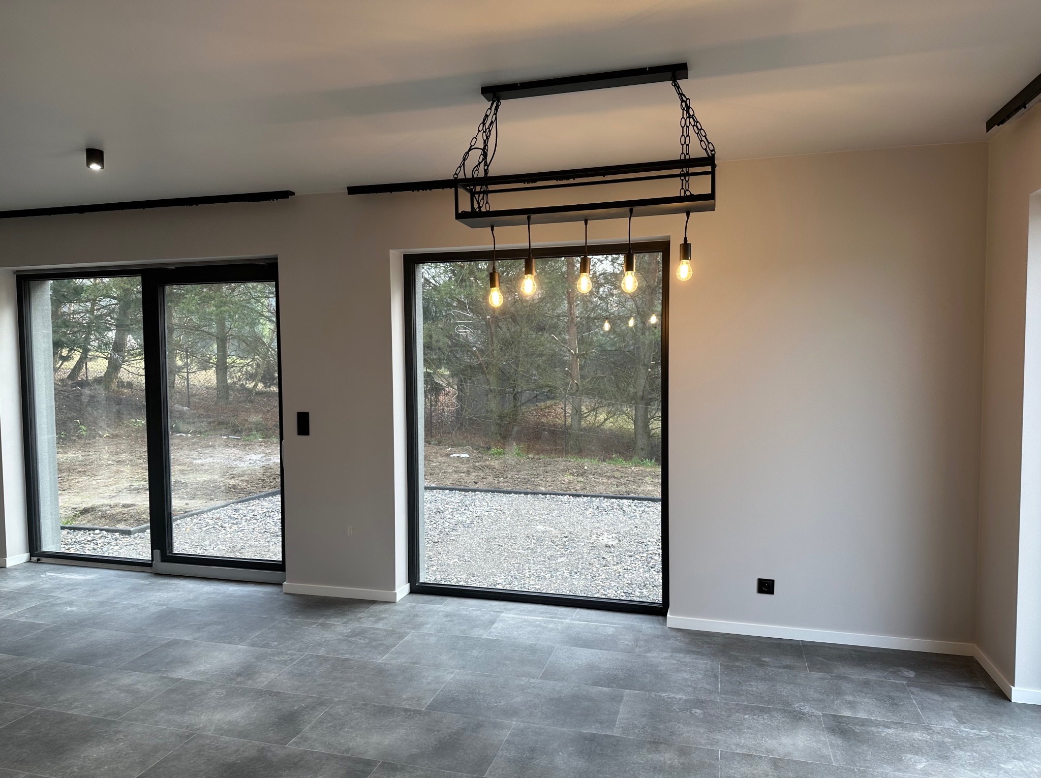Nowoczesne wnętrze z szarymi płytkami podłogowymi i dużymi oknami z czarnymi ramami. Industrialna lampa wisząca z żarówkami Edisona. Świeżo pomalowane ściany w neutralnym odcieniu.
