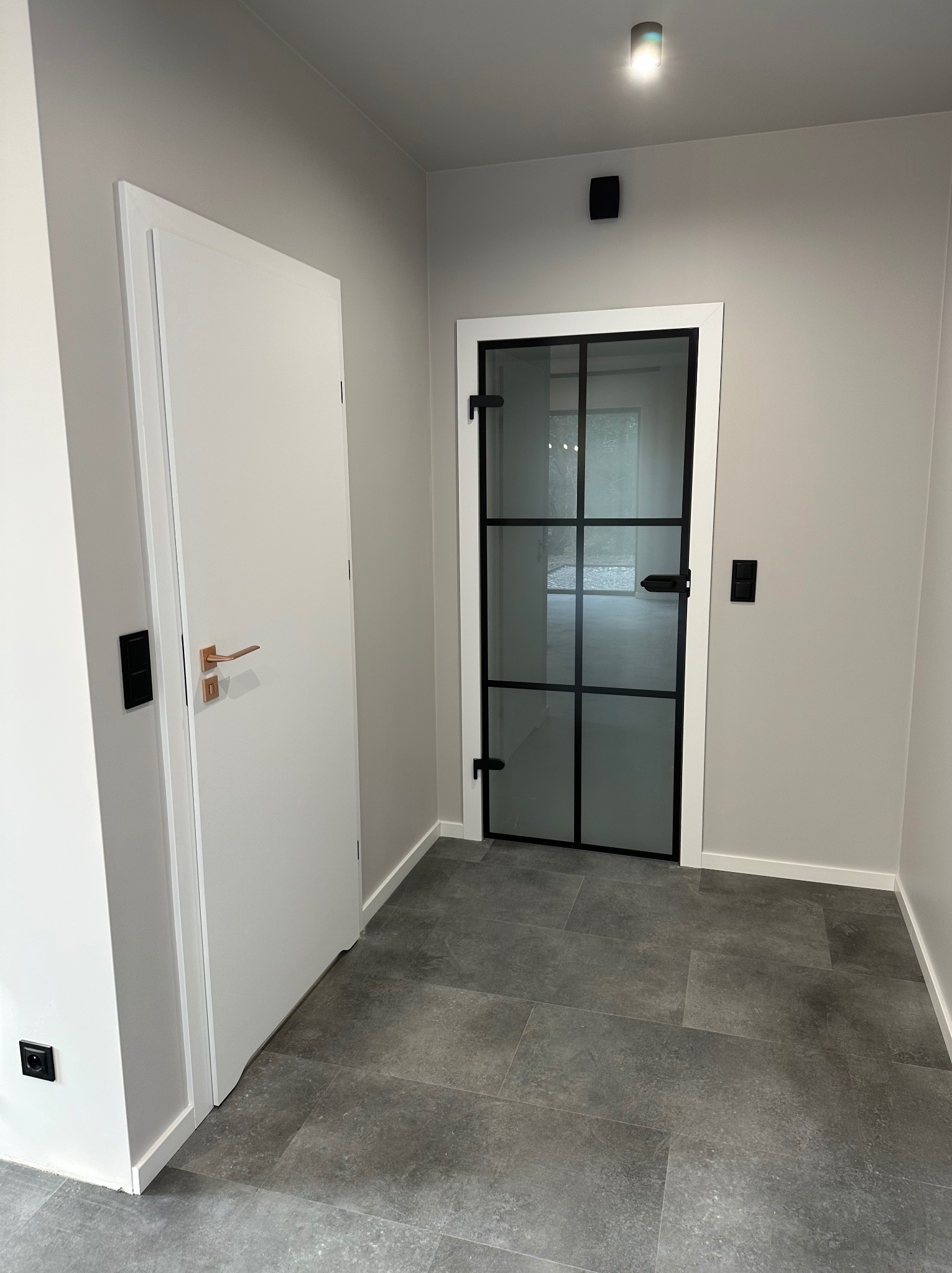 Nowoczesny korytarz z szarą podłogą, białymi drzwiami z miedzianą klamką i przeszklonymi drzwiami z czarnymi szprosami. Minimalistyczne wykończenie, stonowana kolorystyka.