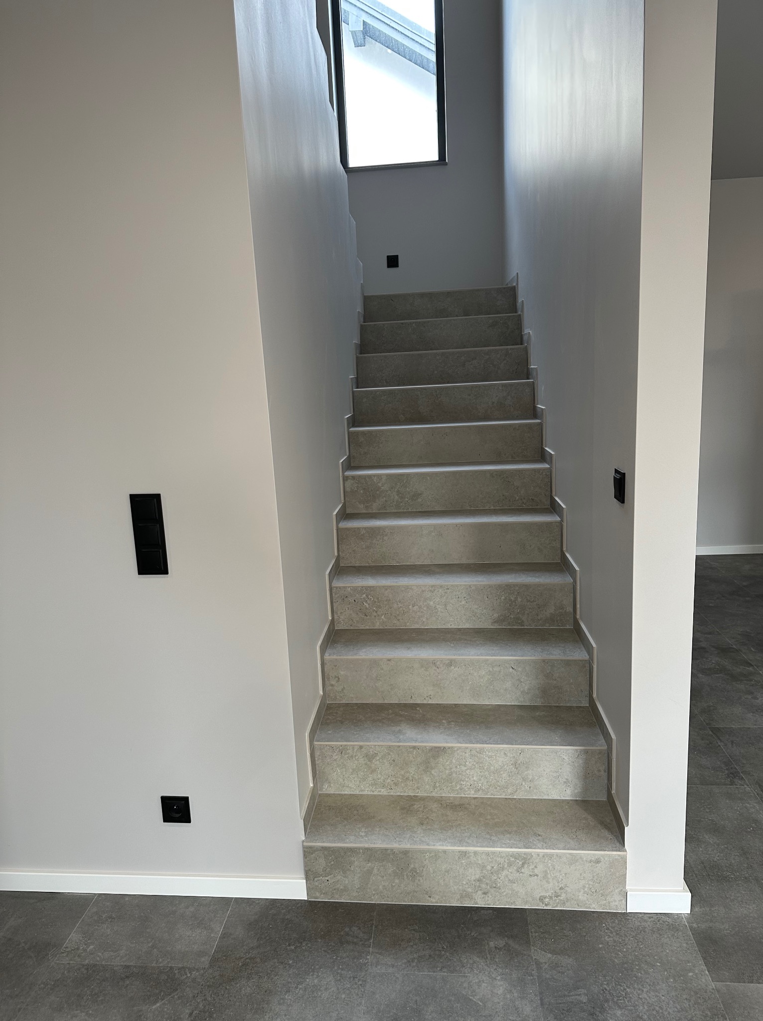 Nowoczesne schody z szarymi stopniami i gładkimi, jasnymi ścianami. Minimalistyczny design z czarnymi włącznikami i szarą podłogą.