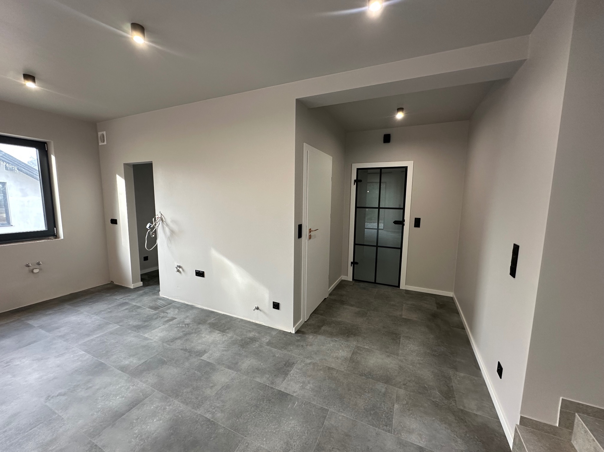 Jasne, wykończone wnętrze z szarą podłogą, świeżo pomalowanymi ścianami i czarnymi akcentami, takimi jak rama drzwi i gniazdka elektryczne. Nowoczesne oświetlenie sufitowe.