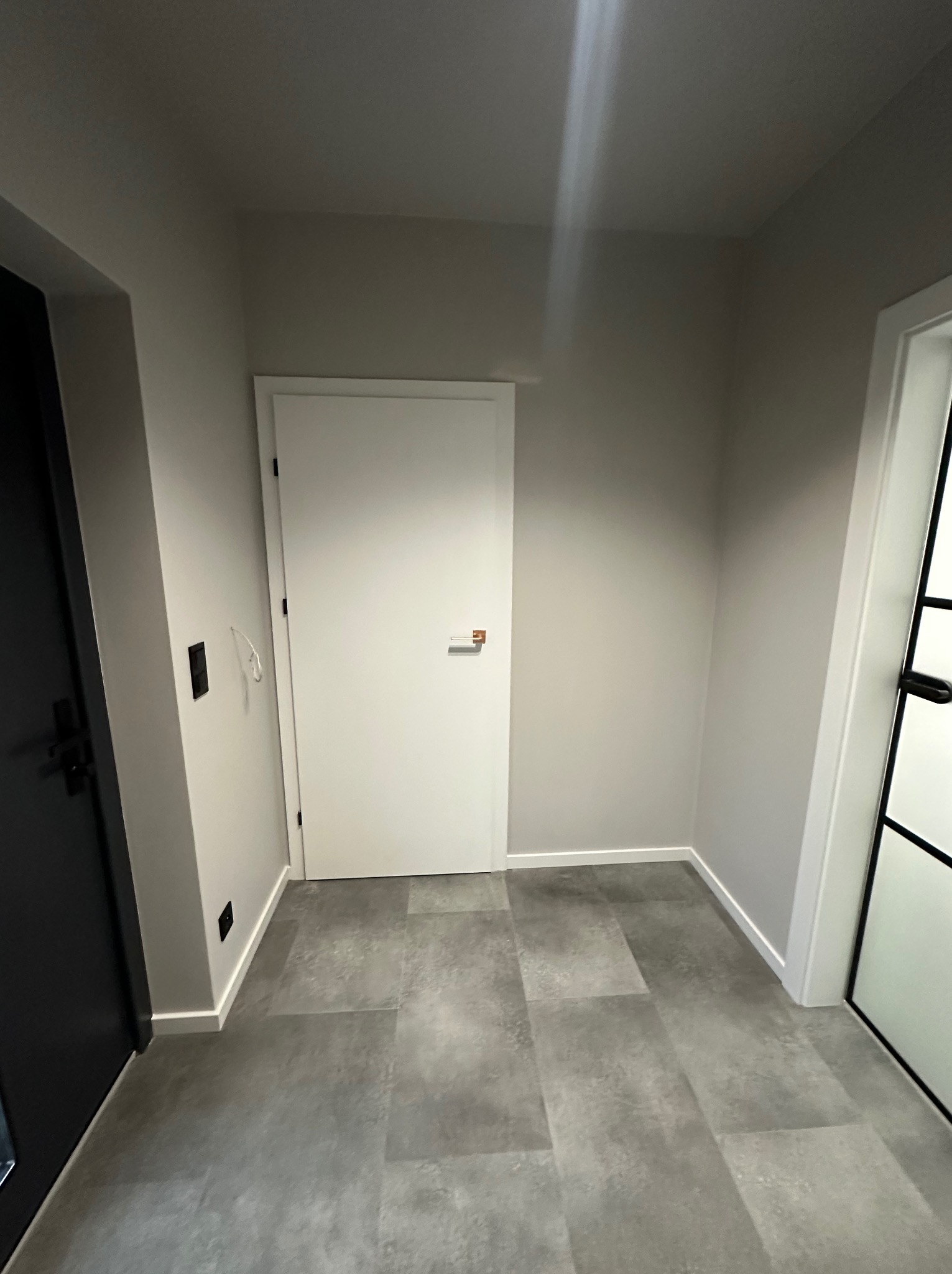 Nowoczesny przedpokój z szarą podłogą imitującą beton, białe drzwi wewnętrzne i minimalistyczne wykończenie ścian. Widoczne detale: listwy przypodłogowe i czarne akcenty.