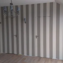 Pokój po malowaniu w szaro-białe pasy z dwiema ukrytymi drzwiami, drewnianą podłogą i dekoracyjnym żyrandolem.