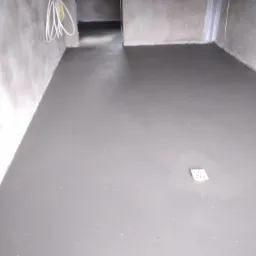 Świeżo wylana, gładka posadzka w surowym wnętrzu z widocznymi przewodami elektrycznymi i kratką ściekową.