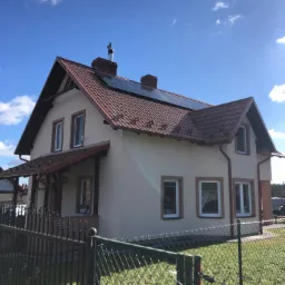 Dom jednorodzinny z panelami słonecznymi na dachu z czerwonej dachówki, widoczny komin z metalowym wkładem, ogrodzenie z siatki i błękitne niebo z chmurami w tle.