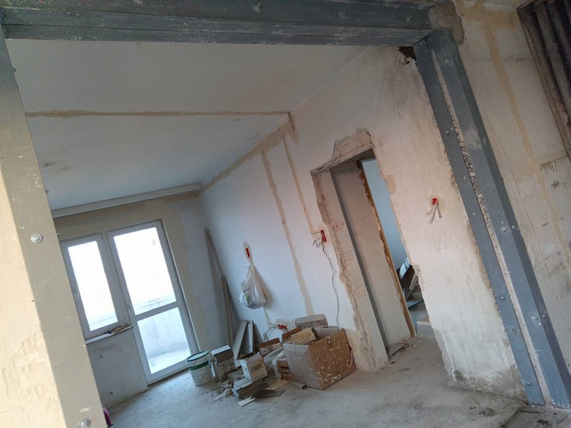 Wnętrze w trakcie remontu, widoczne ślady po usuniętych ścianach, odsłonięte przewody elektryczne, puszki z farbą i kartony na podłodze, wzmocnienia stalowe w otworze drzwiowym.