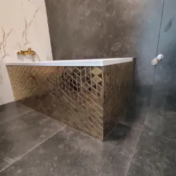 Nowoczesna łazienka z wanną obłożoną złotą mozaiką w geometryczne wzory, ciemnoszara płytka na podłodze i ścianie, armatura w kolorze złota.