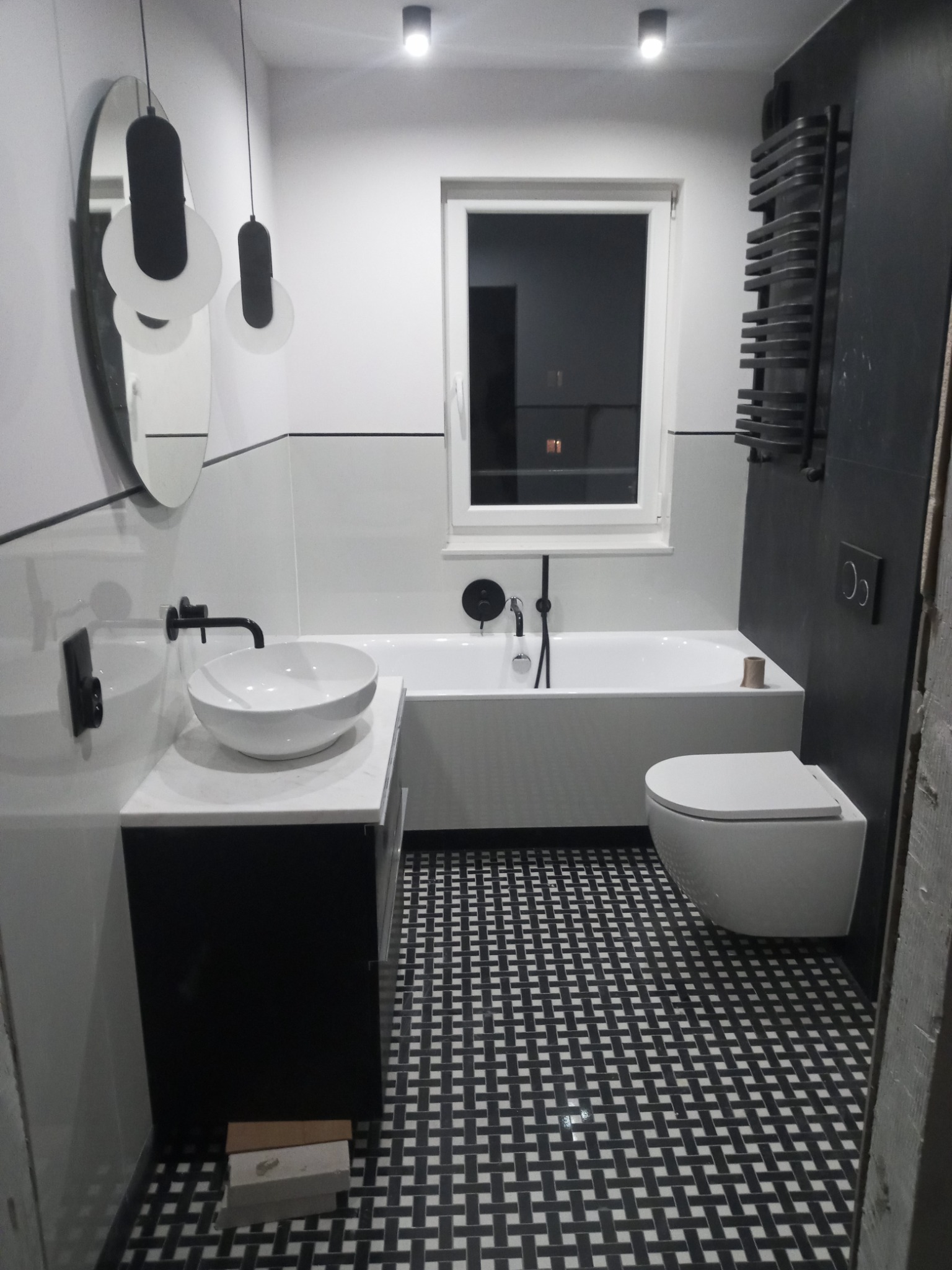 Nowoczesna łazienka w bieli i czerni z geometryczną podłogą, okrągłym lustrem i designerskimi lampami wiszącymi.