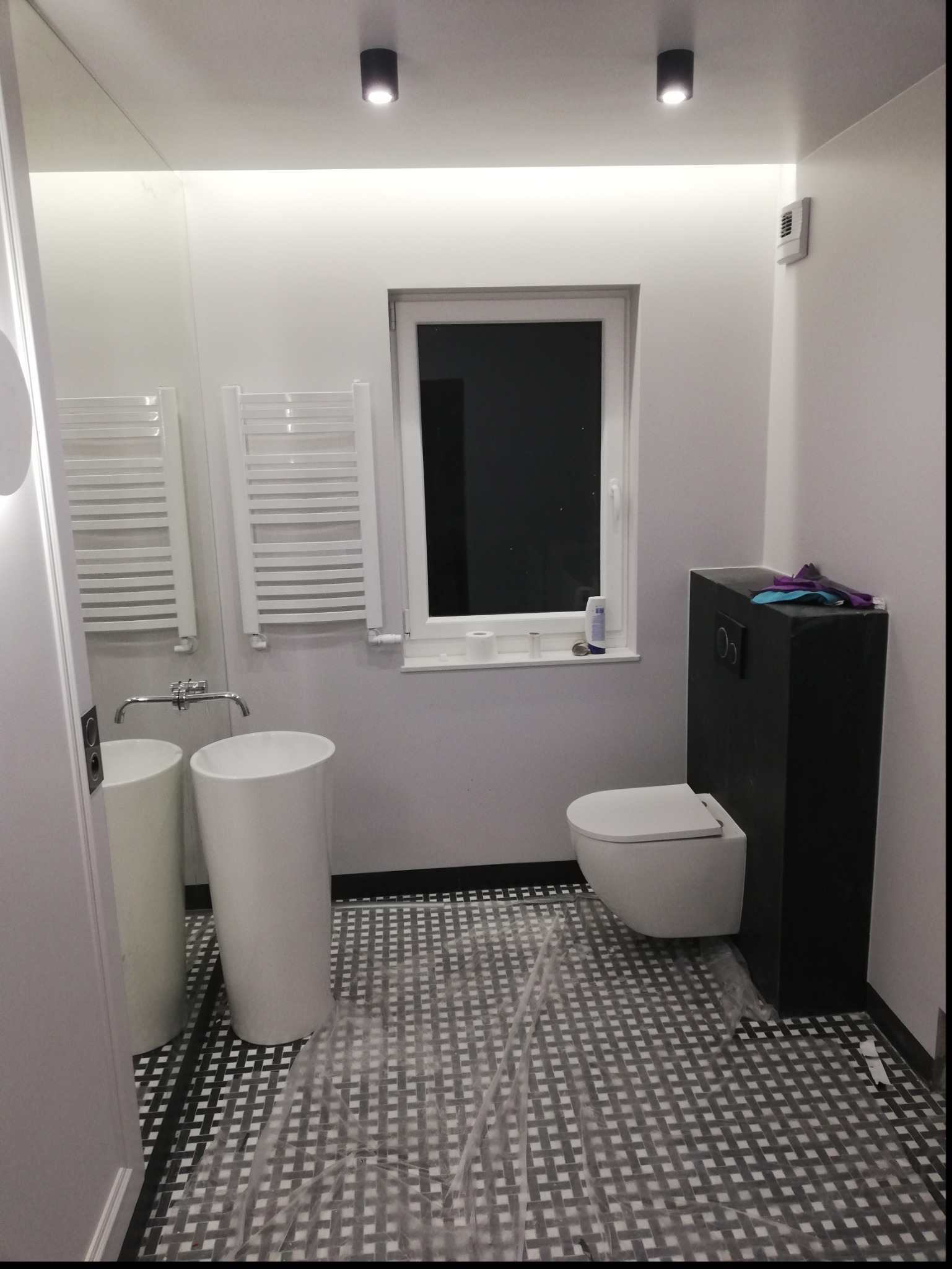 Nowoczesna łazienka w trakcie wykańczania: biała armatura (umywalka i toaleta), czarne akcenty, wzorzysta podłoga zabezpieczona folią ochronną.