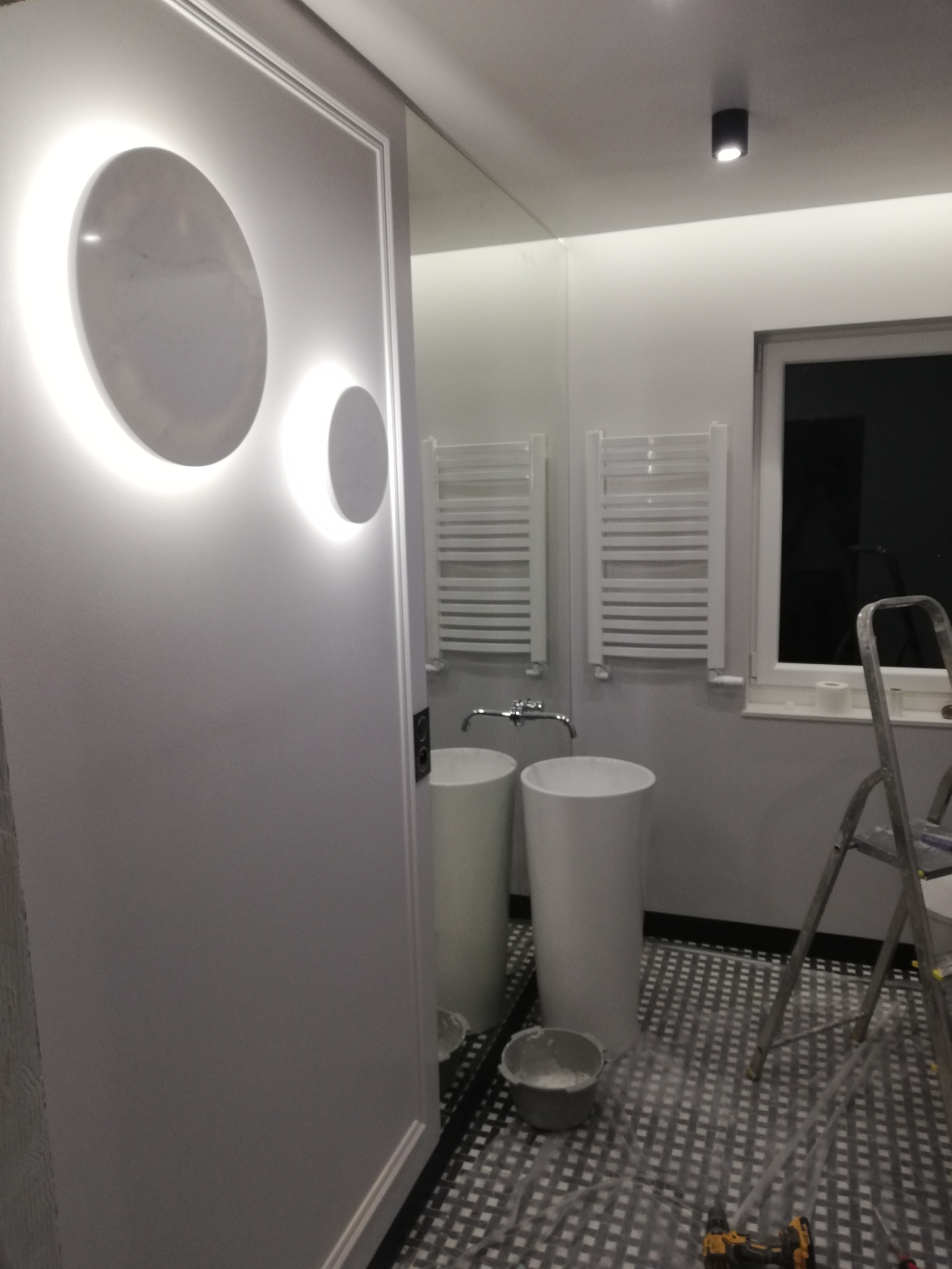 Nowoczesna łazienka w trakcie wykończenia, widoczne dwie białe umywalki na postumentach, grzejniki drabinkowe, drabina malarska i narzędzia na podłodze w czarno-białą szachownicę.