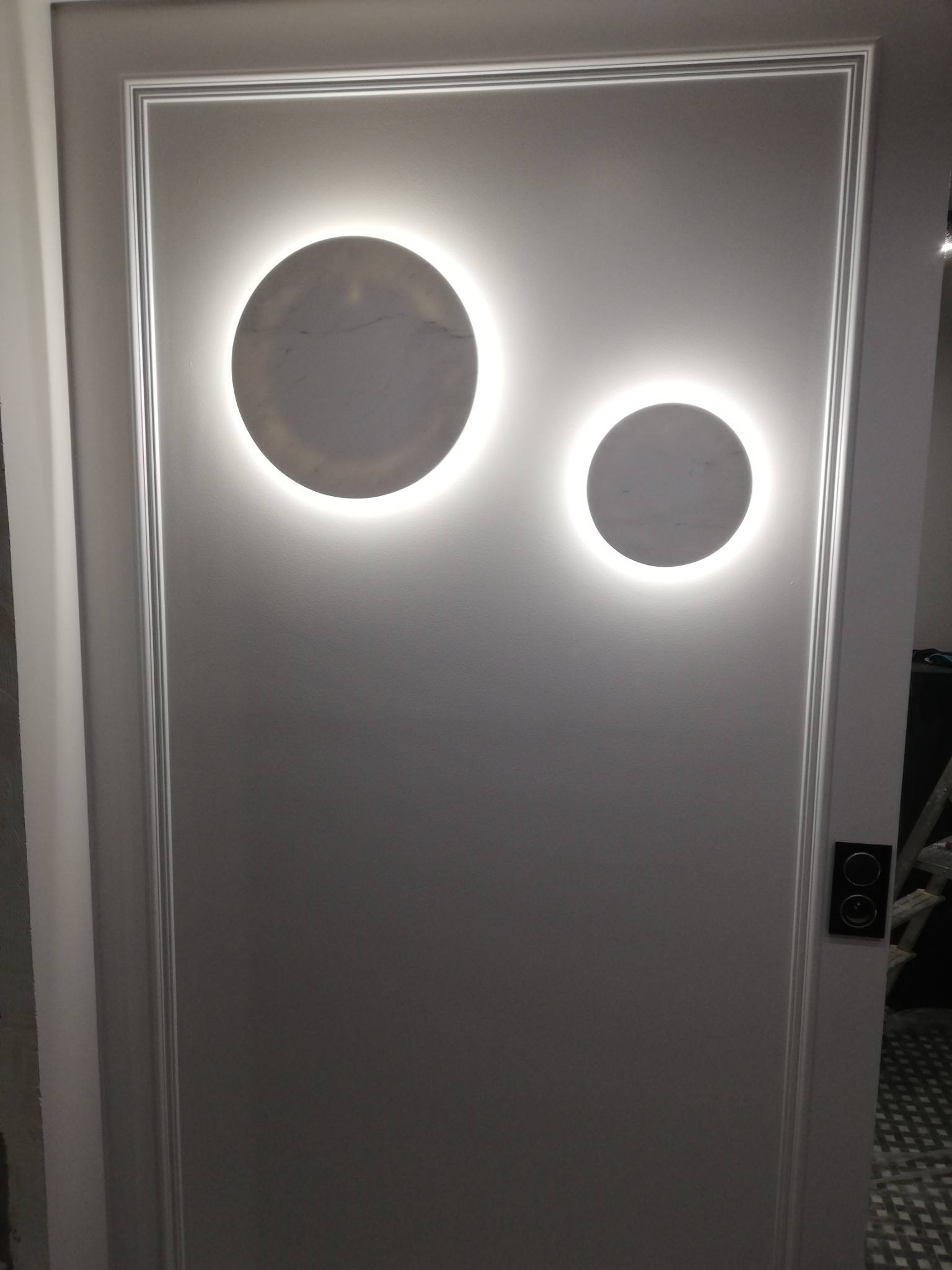 Szare drzwi wewnętrzne z dwoma okrągłymi, podświetlanymi lampami LED o różnej wielkości, umieszczonymi asymetrycznie, ozdobione listwami dekoracyjnymi.