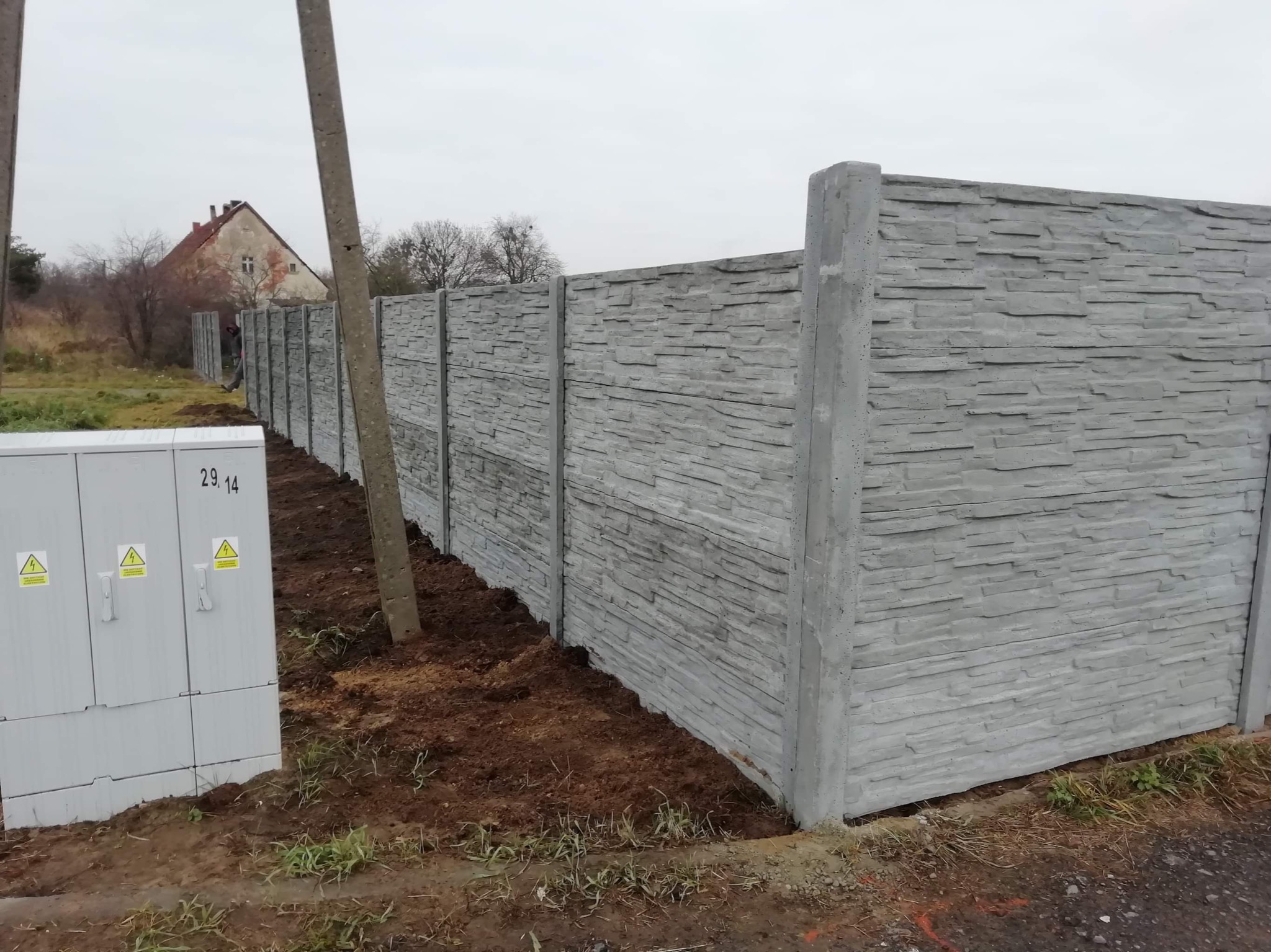 Długi, szary, betonowy płot o wzorze imitującym łupek, z widoczną skrzynką elektryczną i domem w tle, w pochmurny dzień.