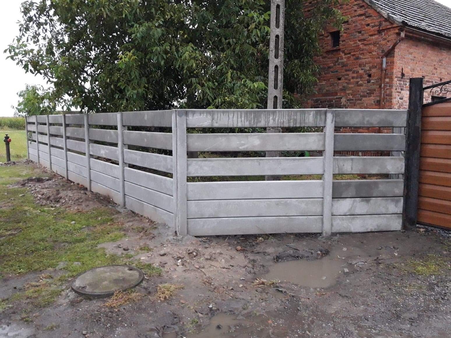 Nowo zamontowane, szare ogrodzenie betonowe z poziomymi panelami i słupkami, widoczne na tle ceglanego budynku i drzew, z hydrantem po lewej stronie i brązową bramą po prawej.