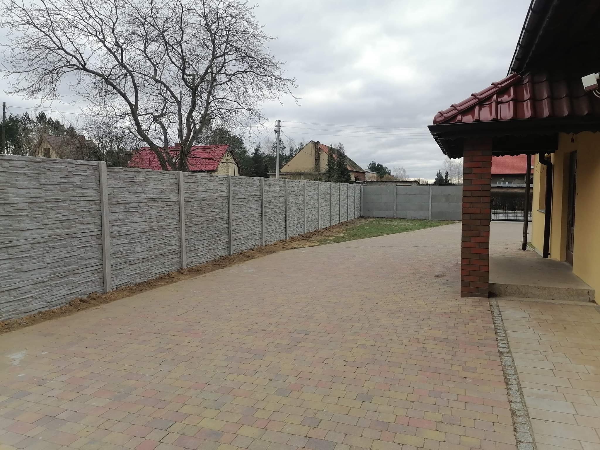 Szary, dwumetrowy płot z paneli betonowych o wzorze imitującym łupek, oddzielający posesję z kostką brukową od sąsiednich domów pod pochmurnym niebem.