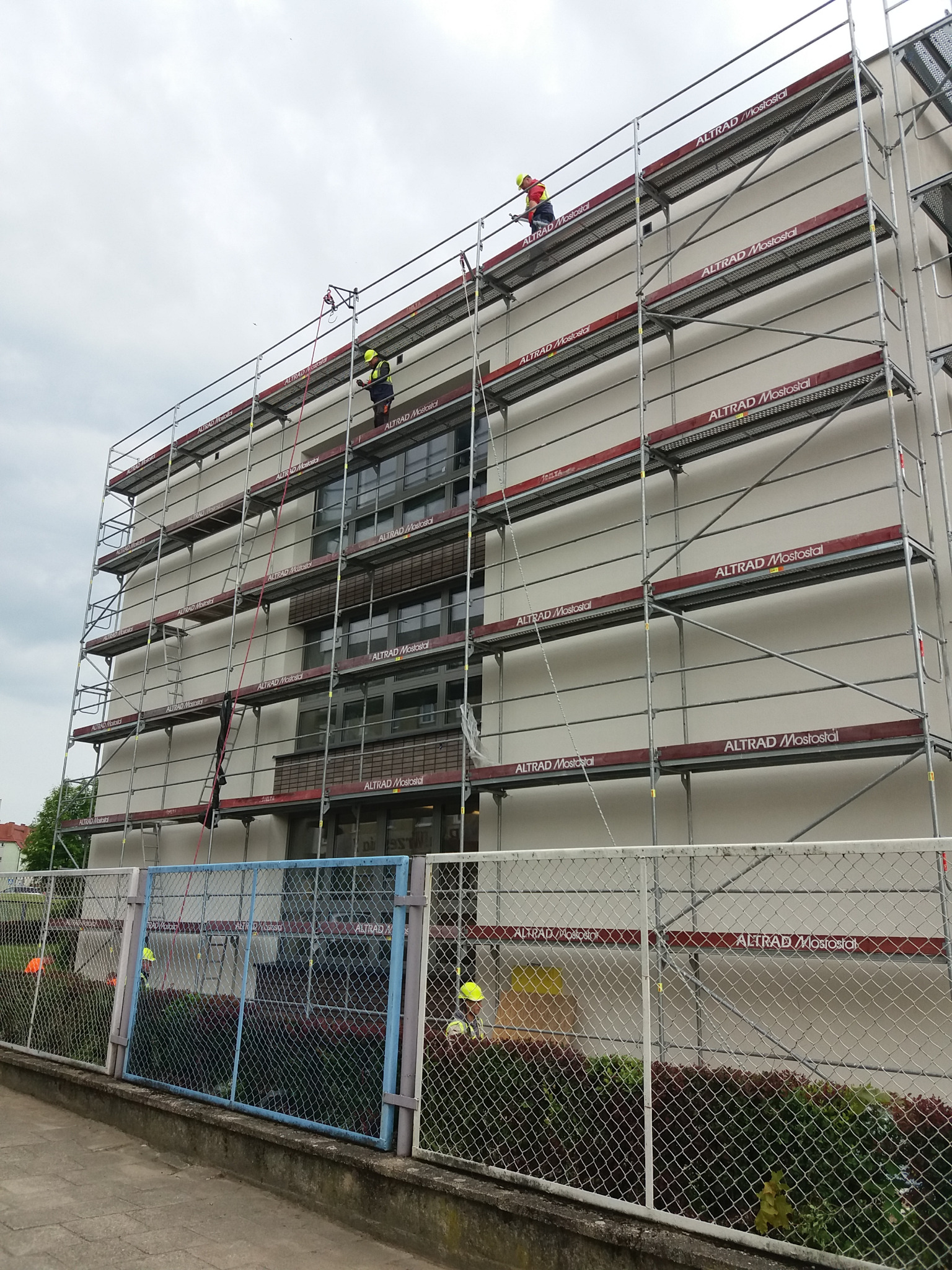 Montaż rusztowania modułowego Altrad Mostostal przy budynku, widoczni pracownicy w kaskach i kamizelkach odblaskowych na różnych poziomach konstrukcji.