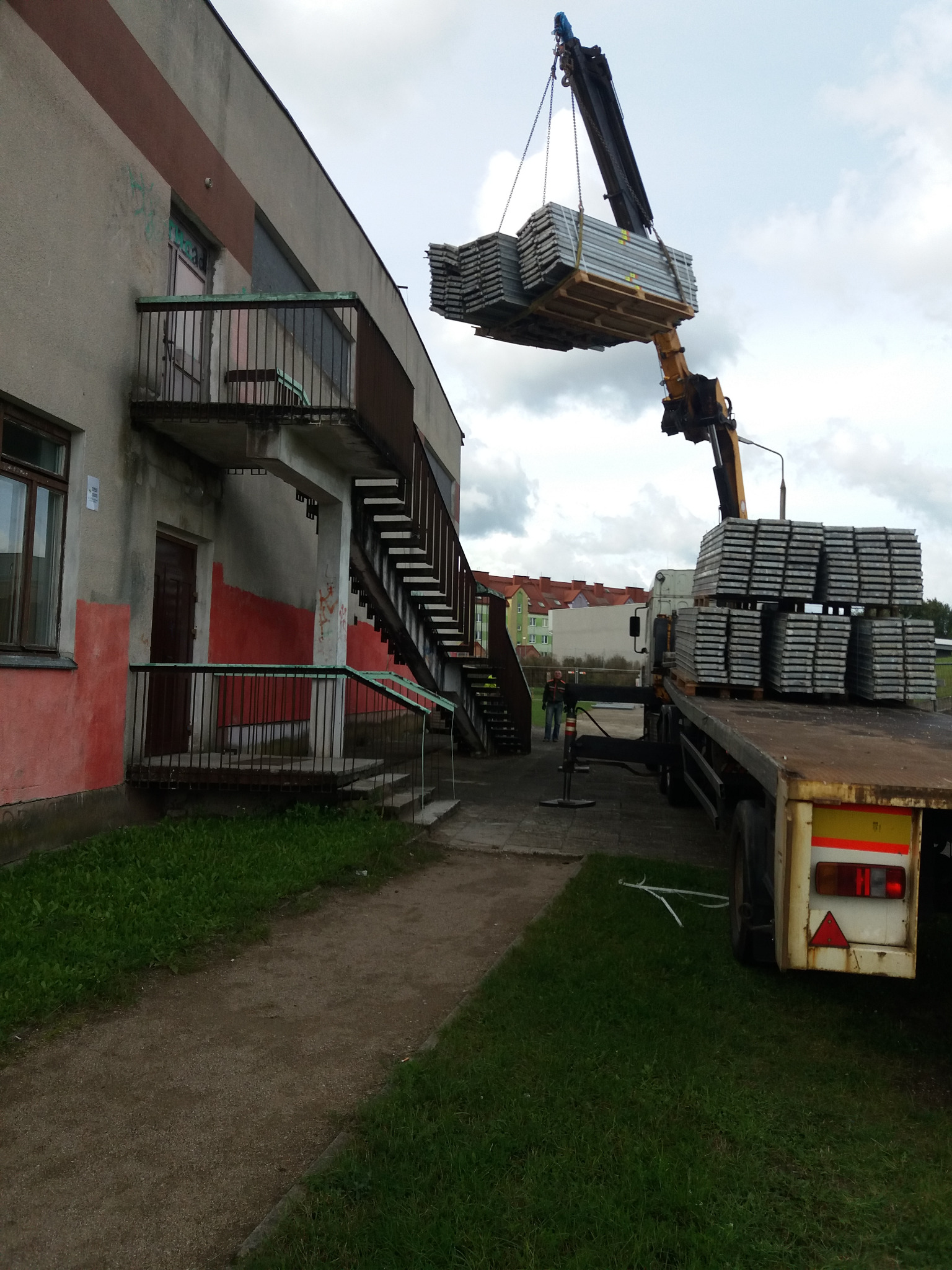 Żuraw samochodowy podnosi paletę z szalunkami na tle budynku z zewnętrznymi schodami i zaparkowanej ciężarówki.