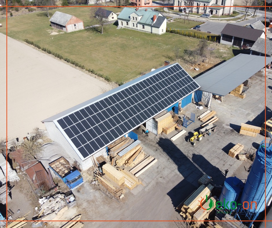Widok z lotu ptaka na duży budynek z panelami słonecznymi na dachu, obok składowane drewno i niebieski silos, w tle zabudowania wiejskie.