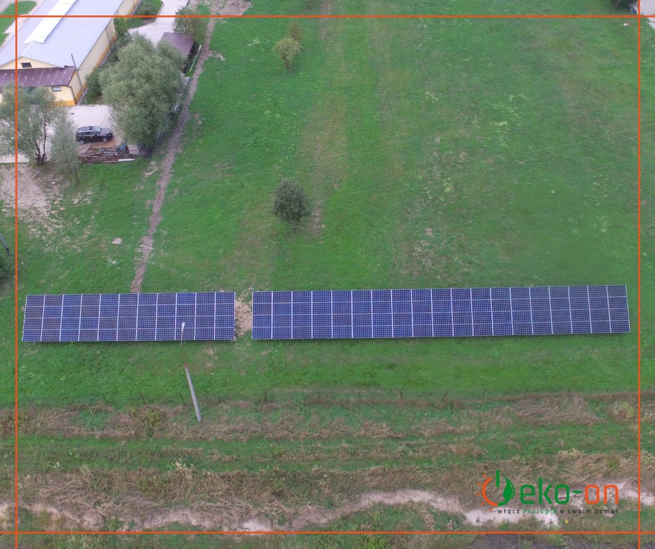 Widok z góry na dwie rzędy paneli słonecznych na zielonym trawniku, obok budynek z drzewami i samochodem, w oddali logo firmy.