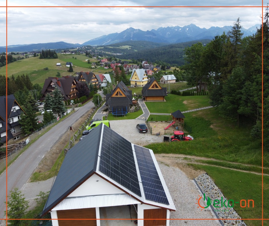 Widok z góry na garaż z panelami słonecznymi na dachu, w tle zabudowa wiejska, góry i las.